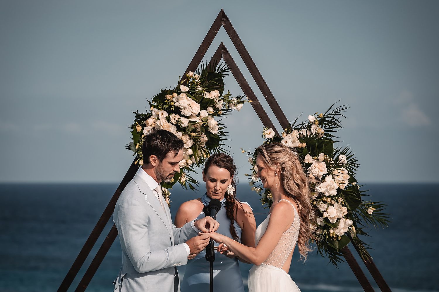 Intimate vow exchange moment under triangular floral arch with ocean view.