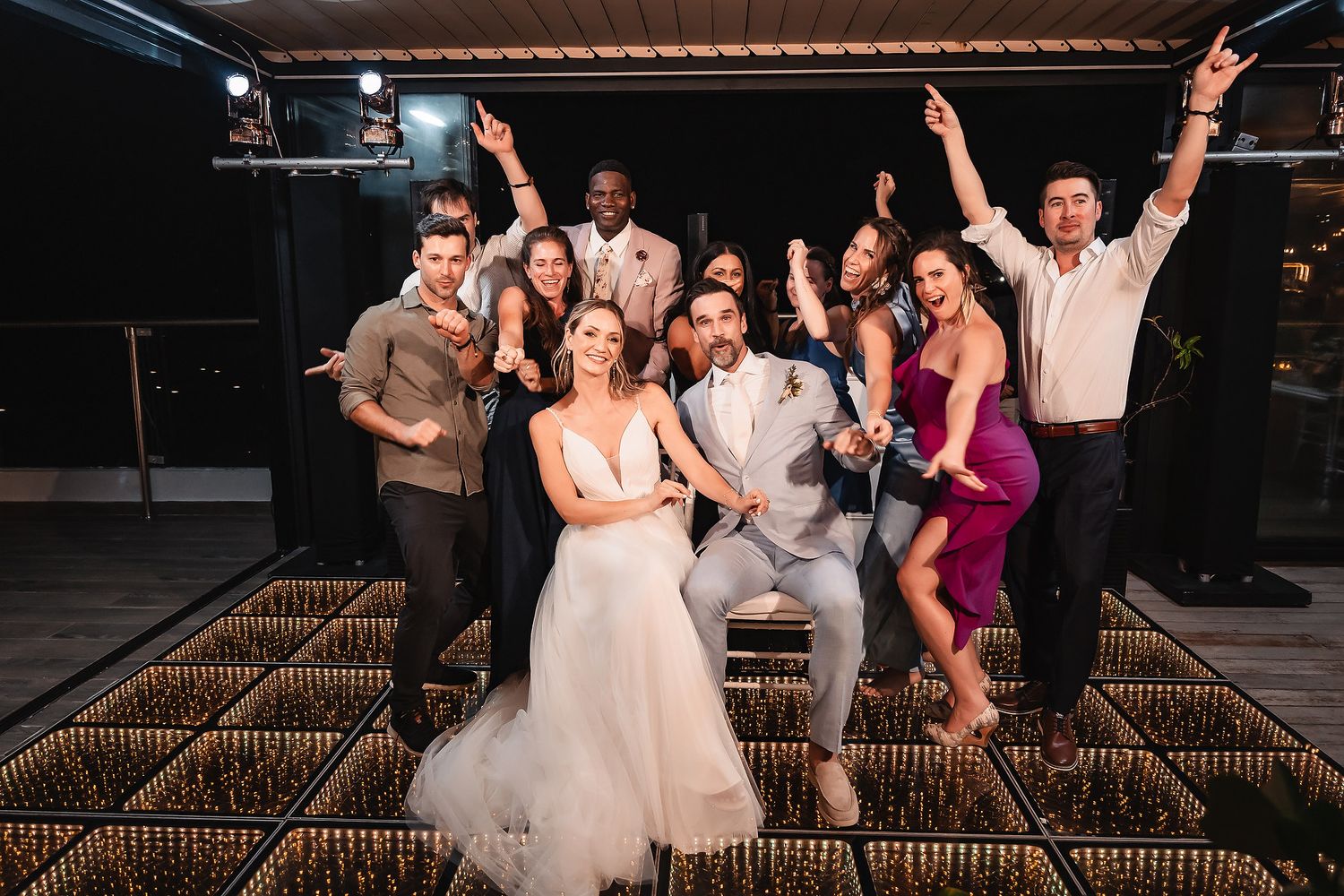 Wedding party poses energetically on illuminated dance floor during evening celebration.