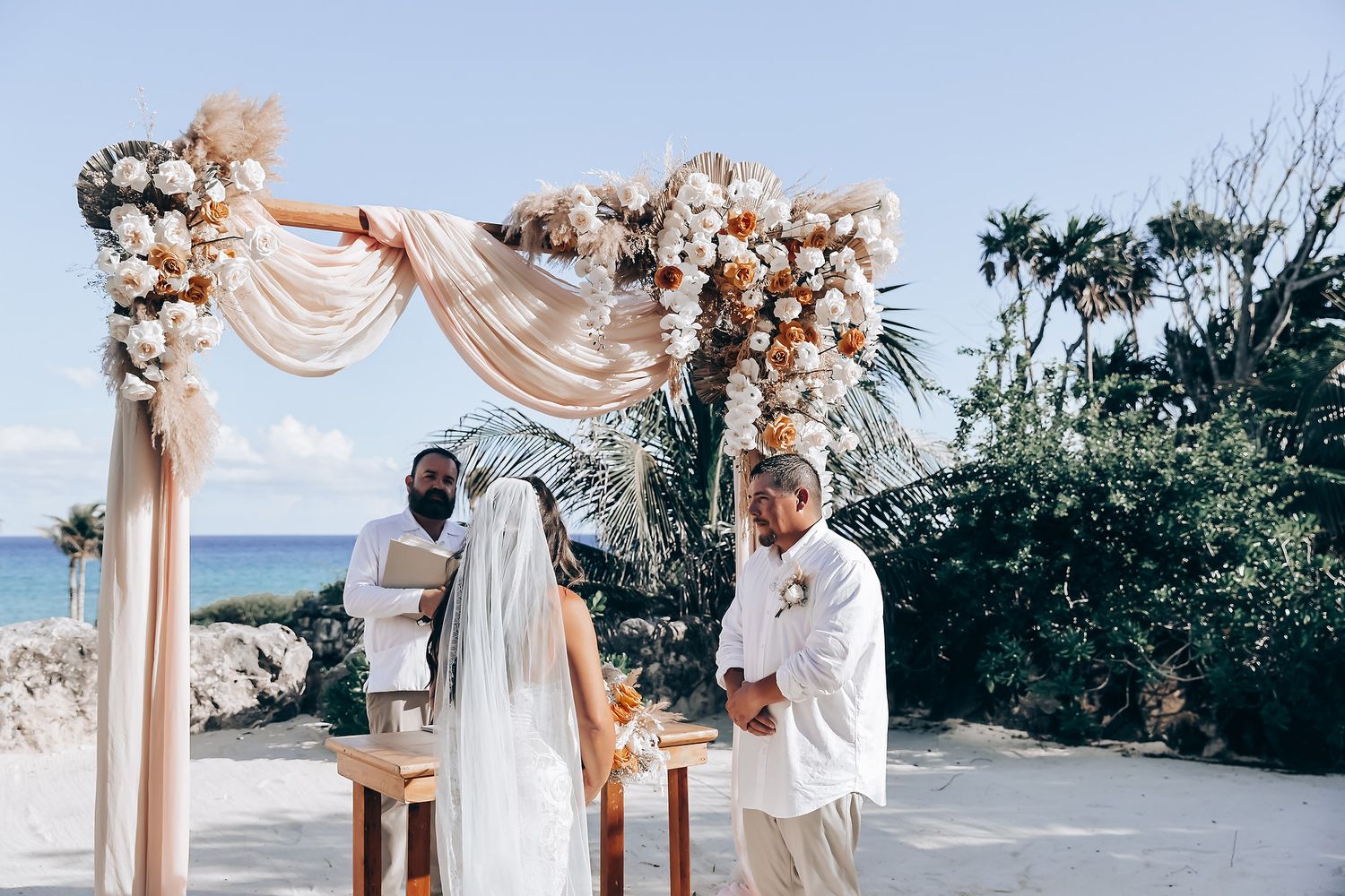 A beachside wedding ceremony setup with a floral arch and draped fabric overlooking the ocean.