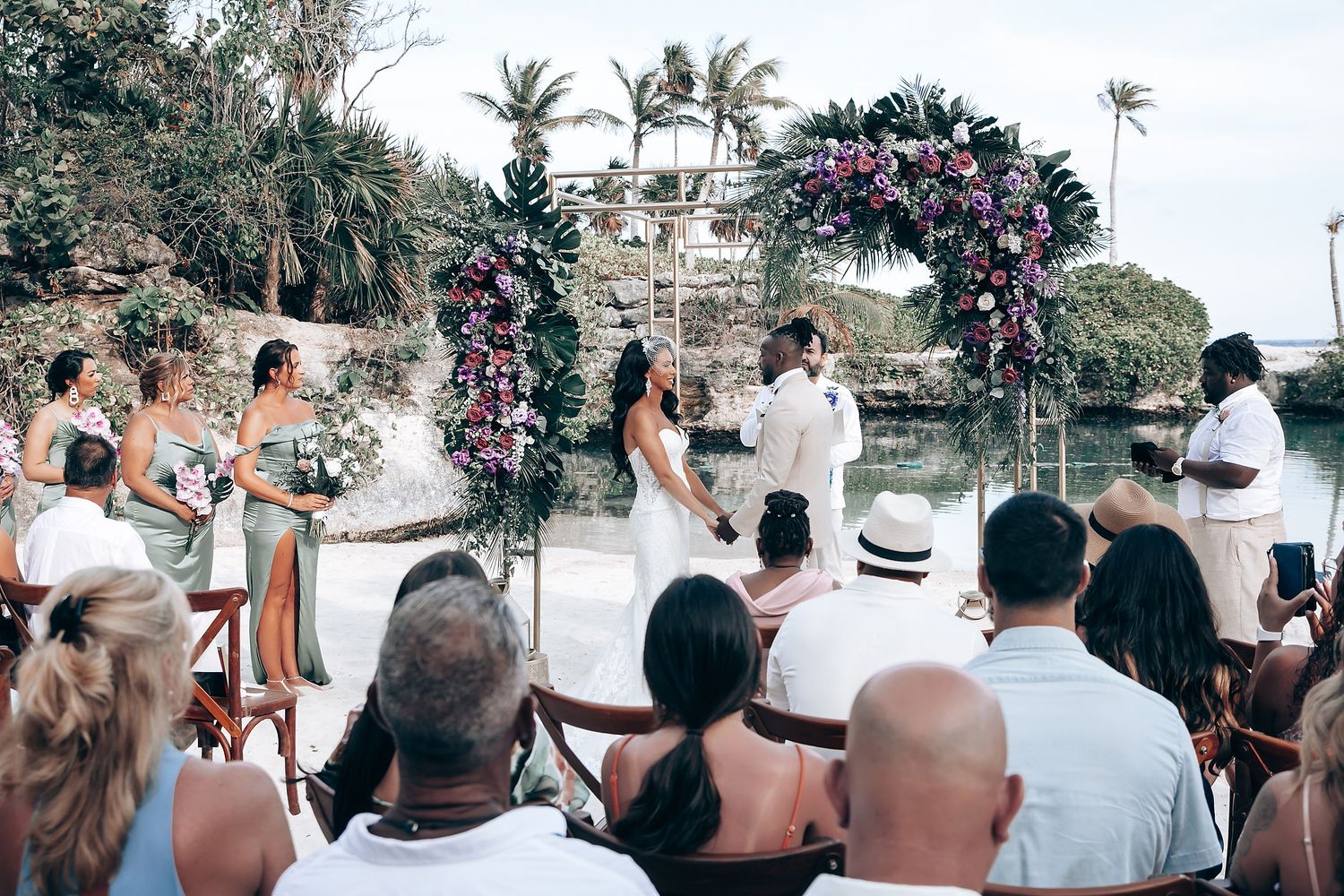 Wedding attendees watching ceremony from wooden chairs at tropical waterfront location.