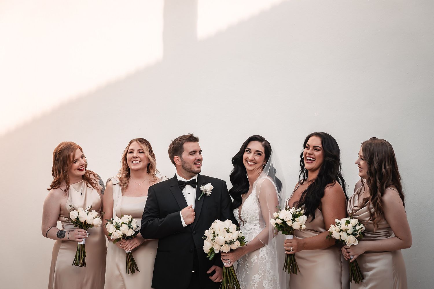 Wedding party in formal attire poses together holding white floral bouquets against a light background.