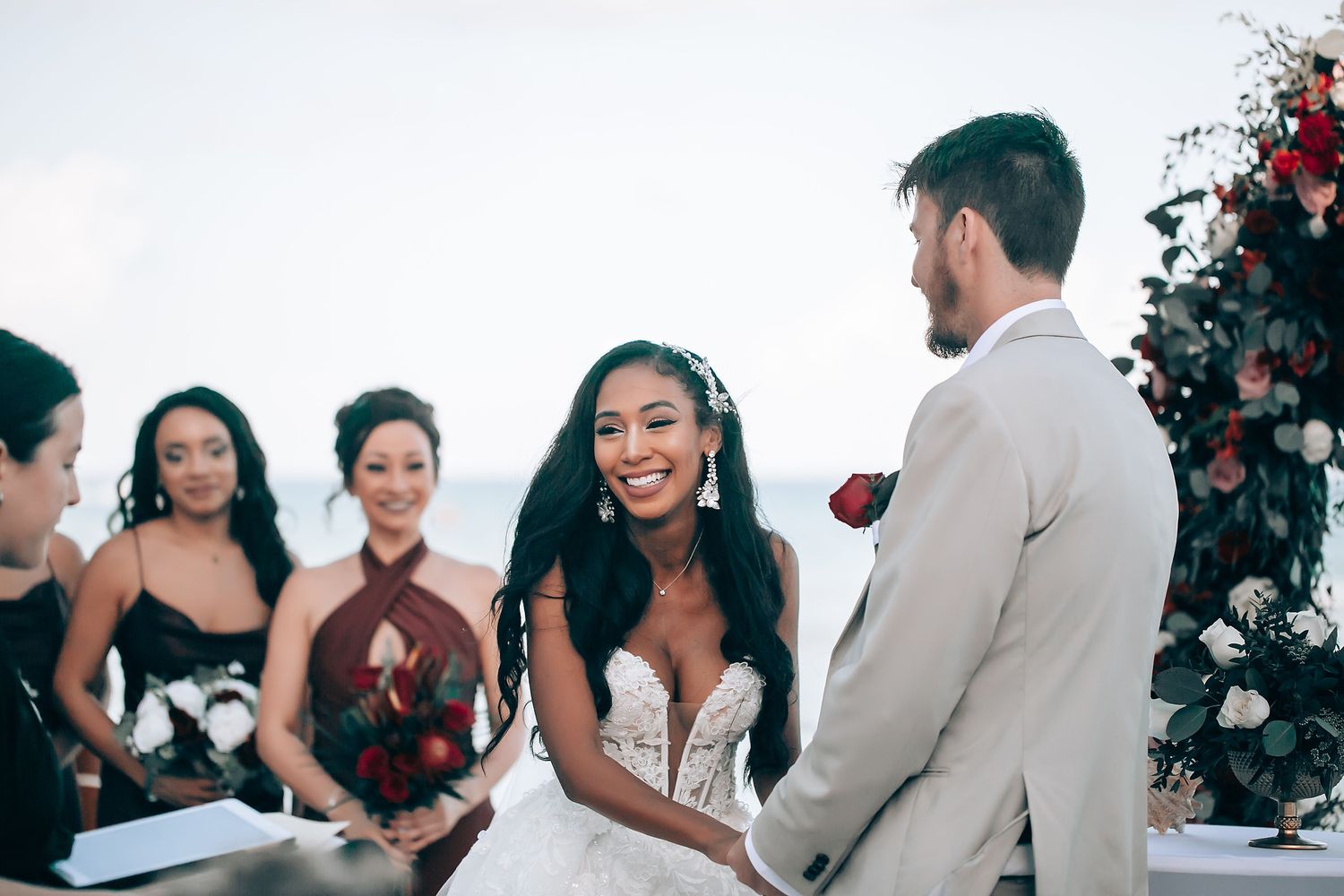 Wedding party celebrating on a beach with burgundy dresses and white decor.