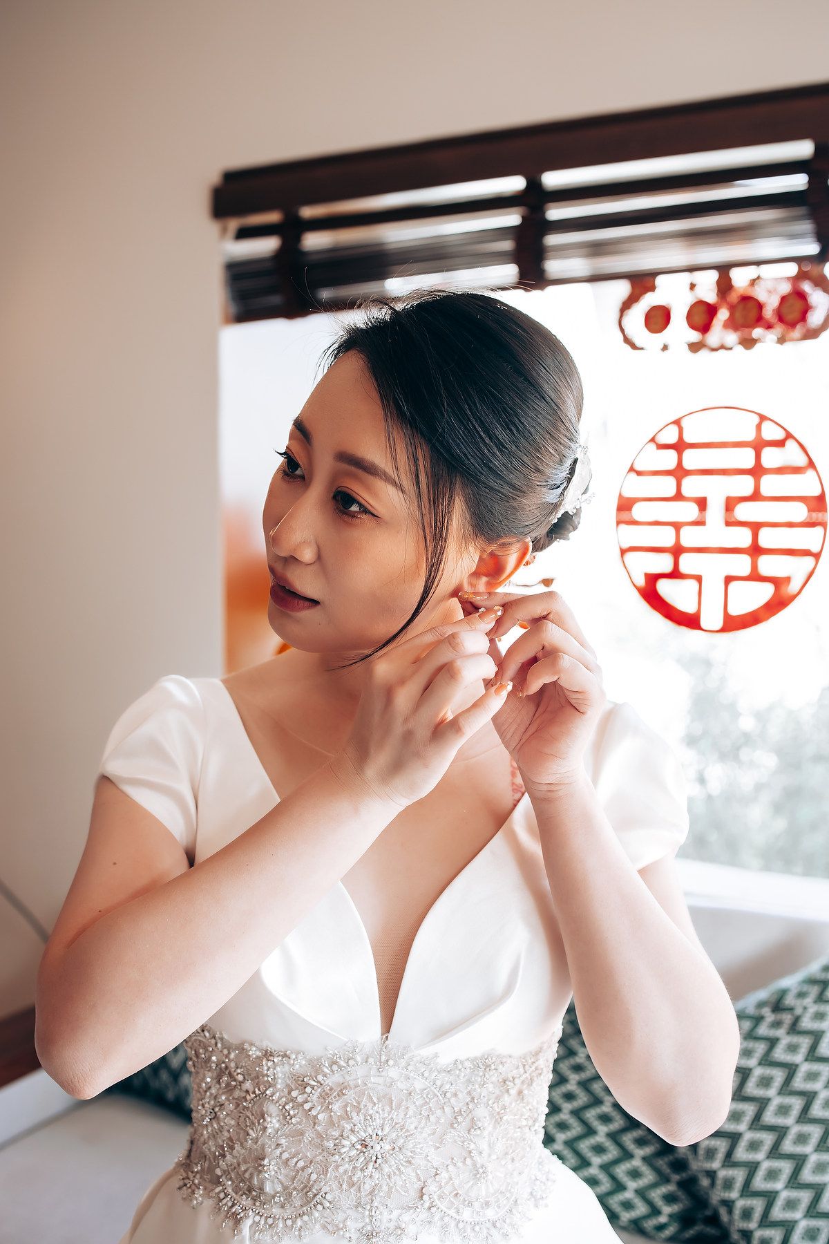 A bride in a white beaded dress puts on earrings while getting ready, with a traditional Chinese symbol visible.