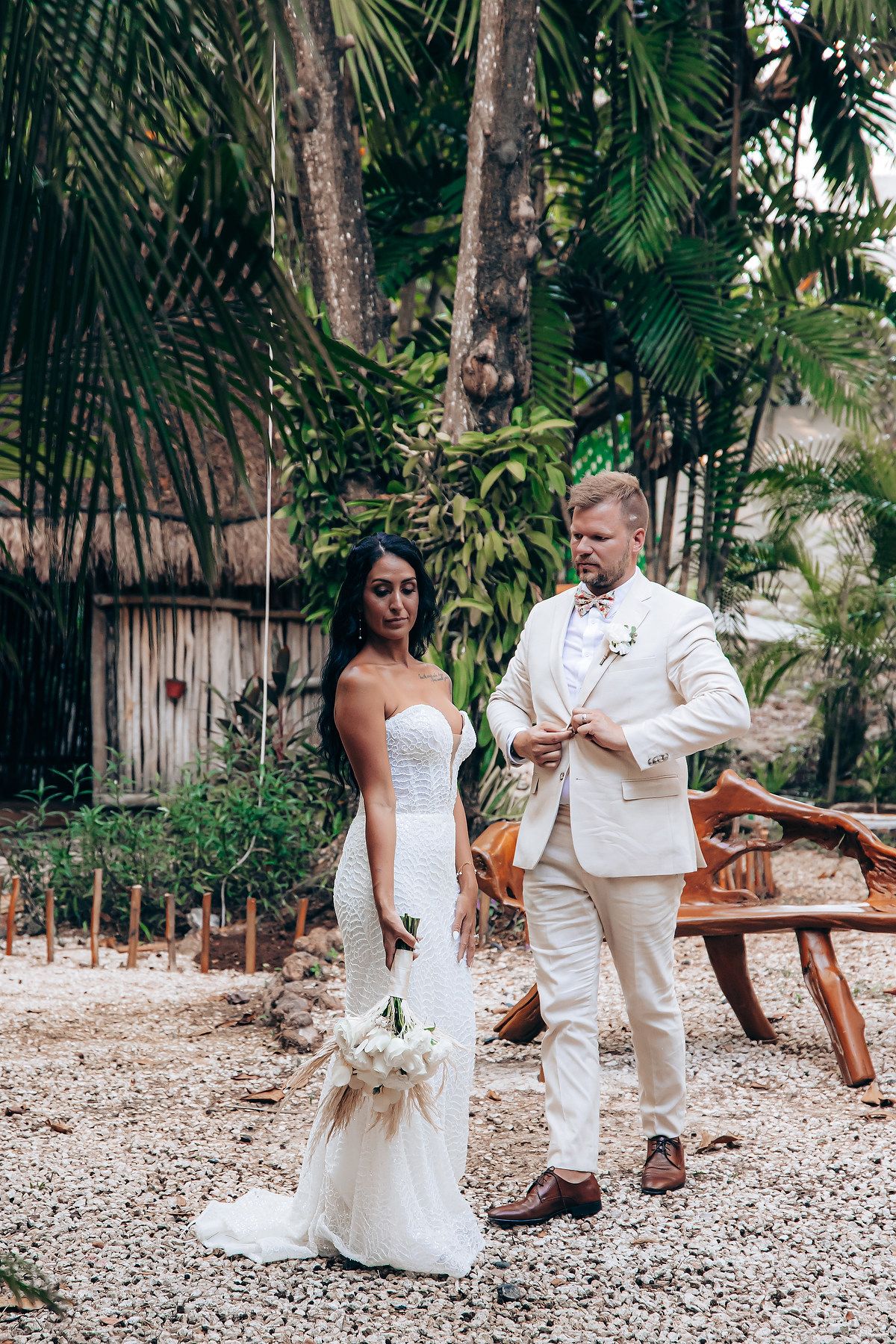 Wedding couple in white attire walking through a tropical garden pathway.