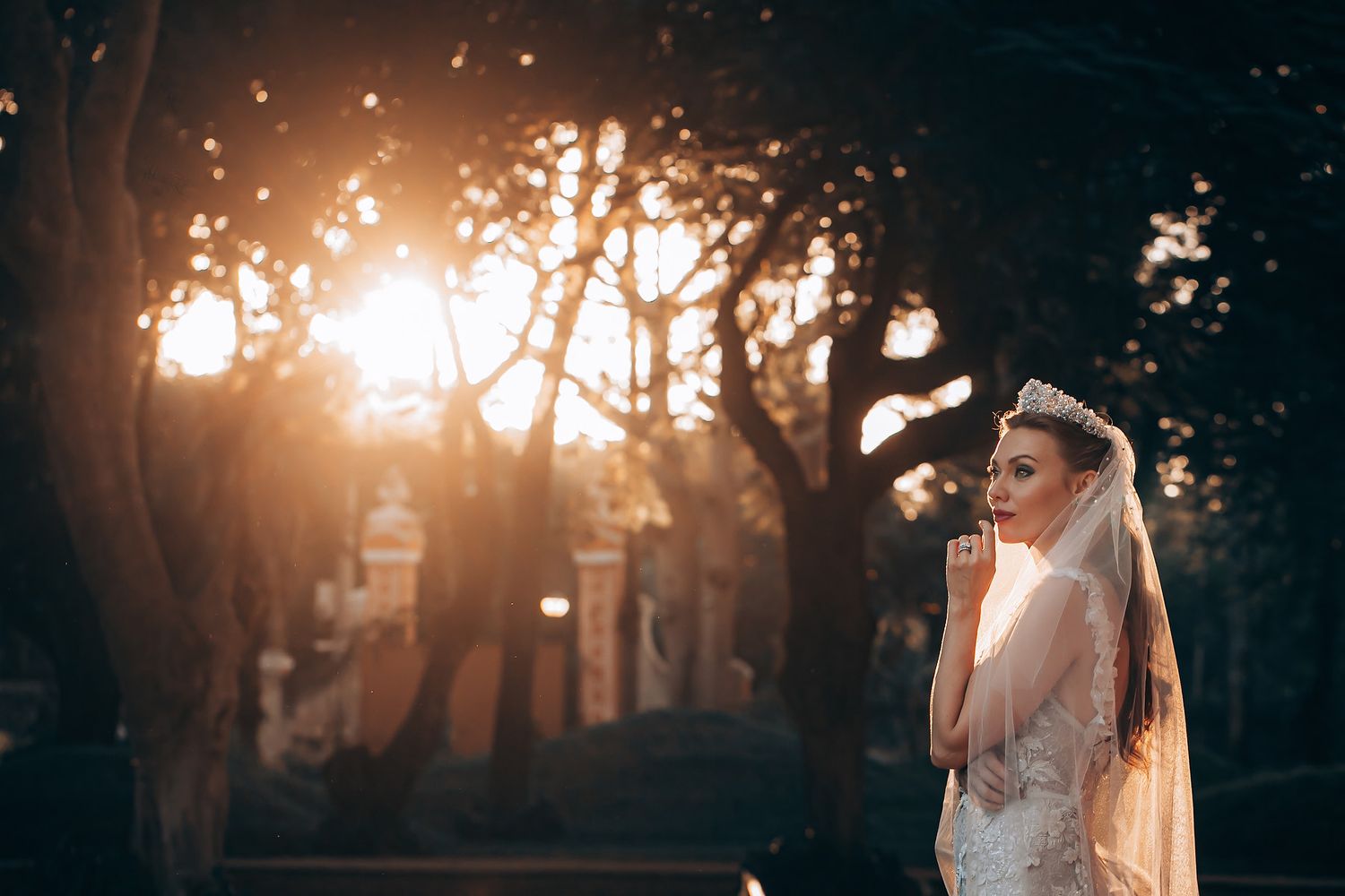 A bride in a white wedding dress poses in a forest backlit by golden sunset rays streaming through the trees.