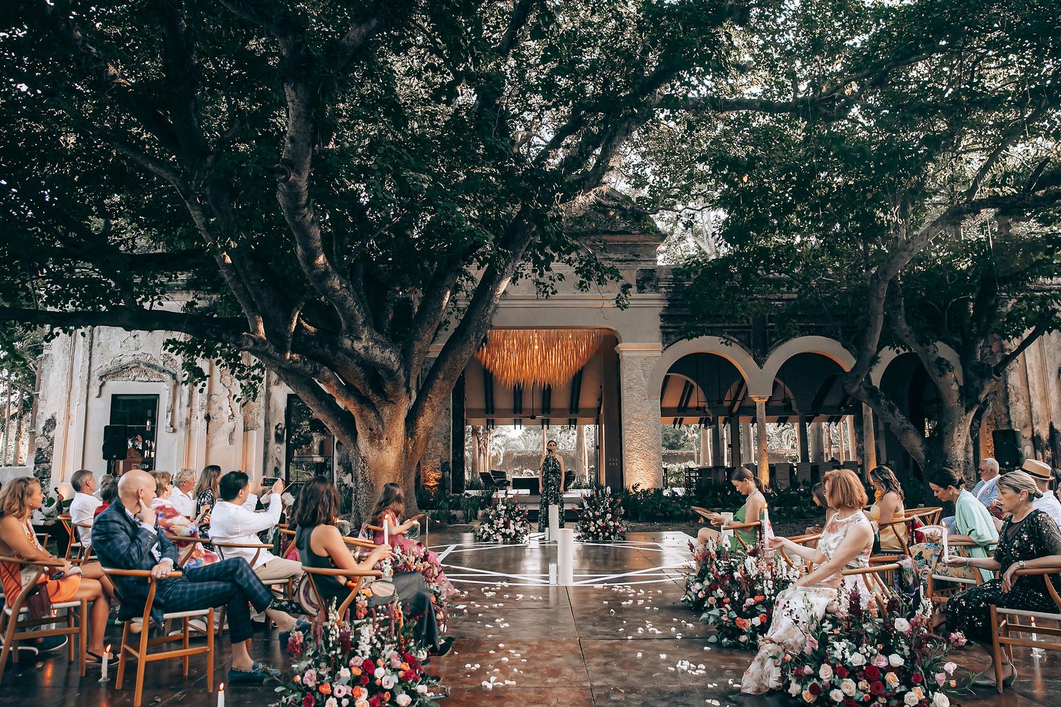 People enjoying a summer day at a crowded outdoor cafe with tables under shady trees in an urban setting.