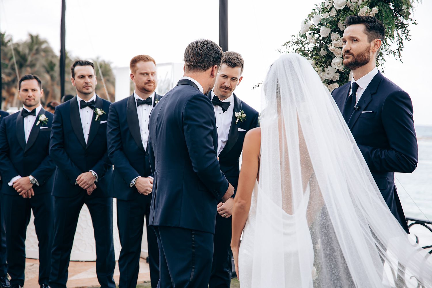 Wedding party in formal black suits and dresses poses together outdoors on a sunny day.