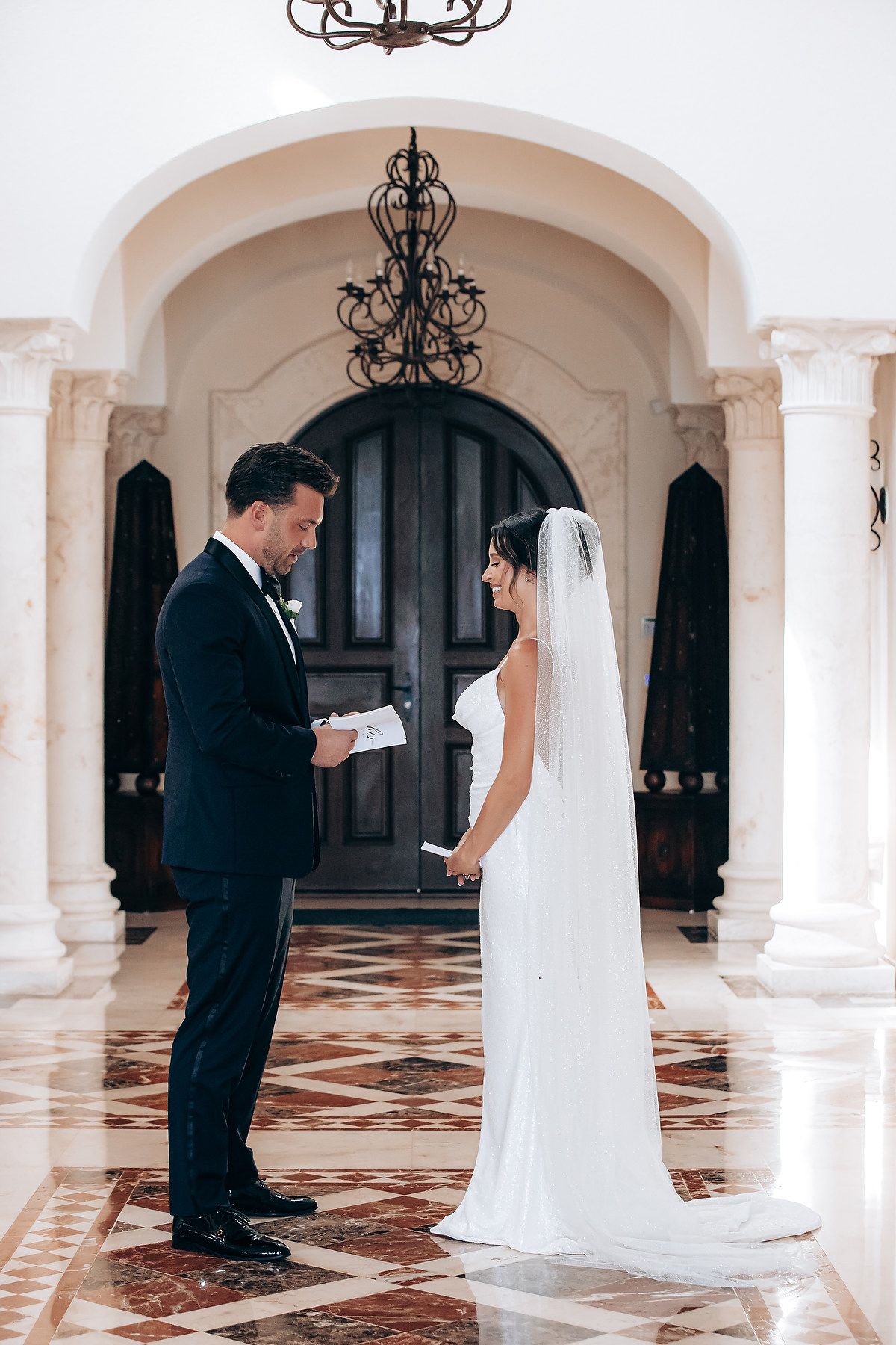 A romantic first look moment between a couple in formal wedding attire under elegant white archways with black chandeliers.