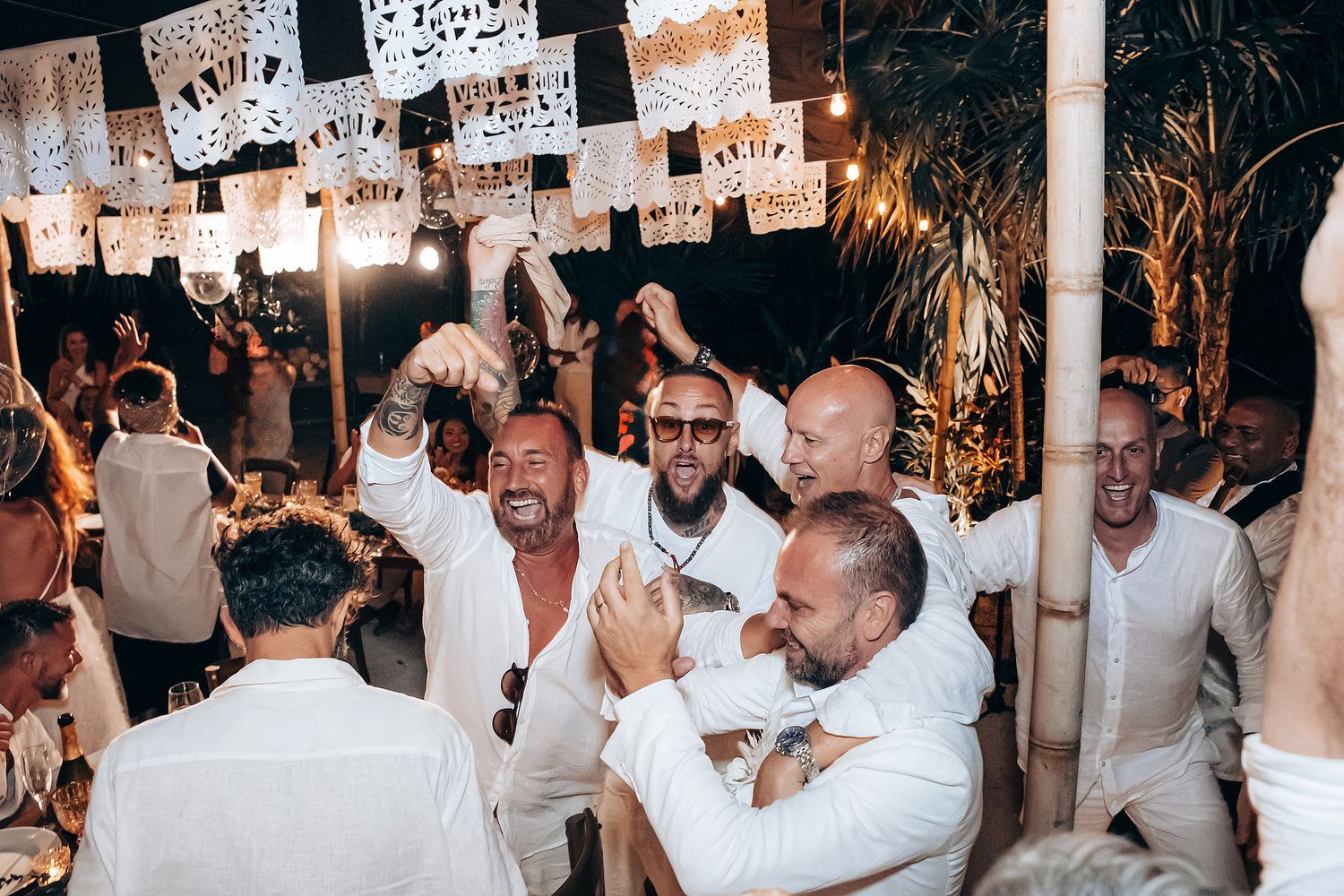 Large group of party guests in white clothing dancing and celebrating at a dimly lit indoor event.