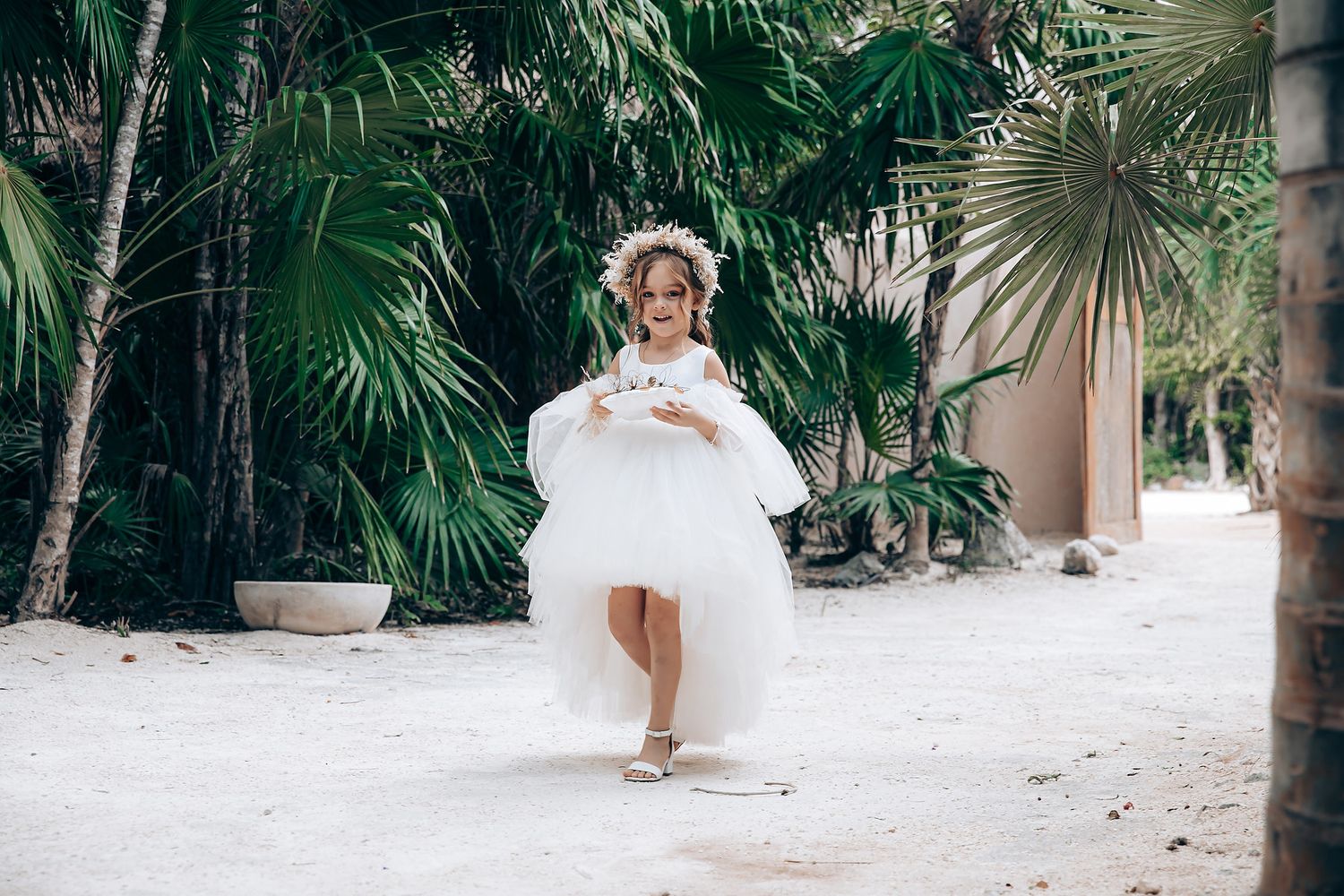 Wedding party members in white dresses walk along a sandy tropical beach path lined with palm trees.