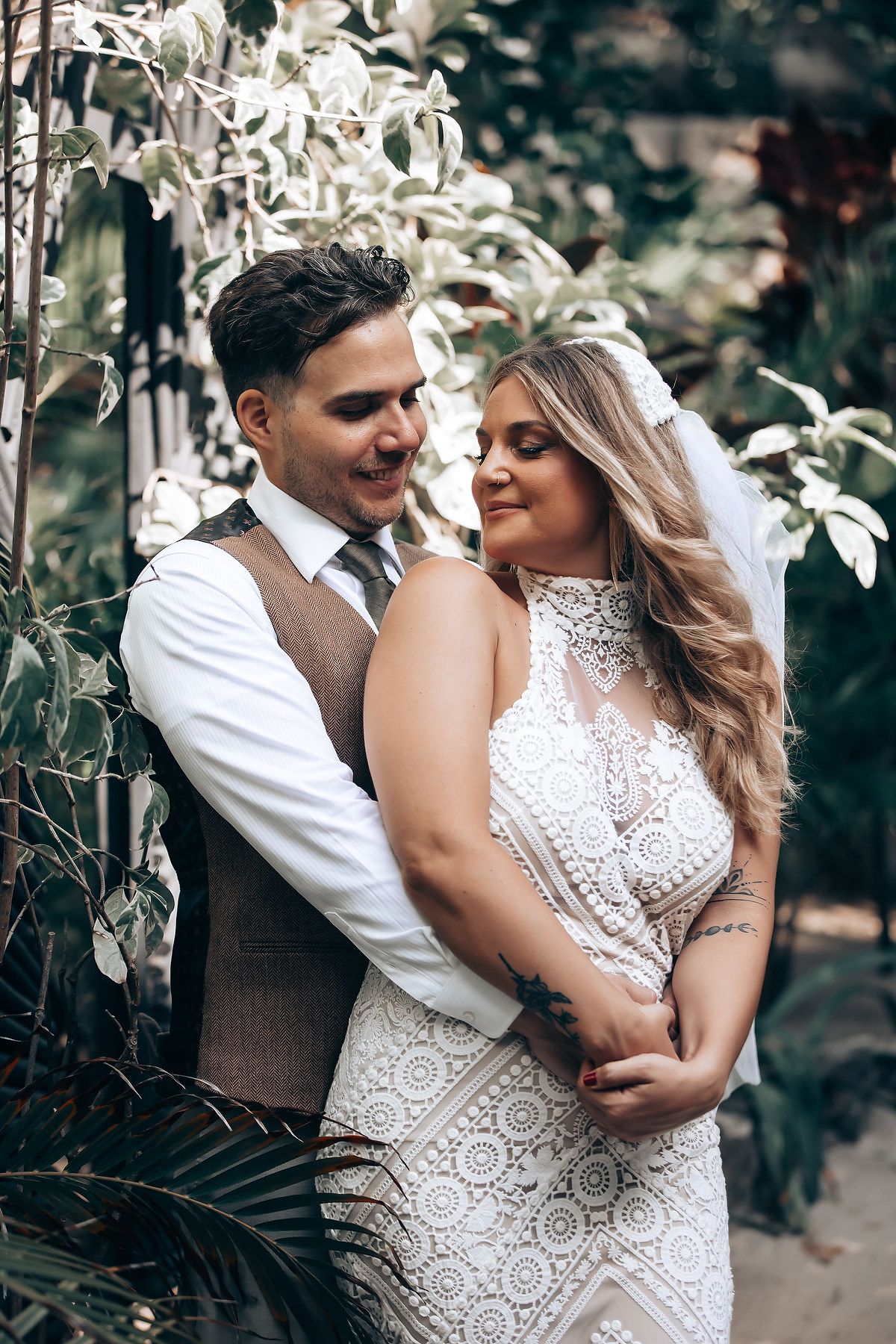 A romantic couple embraces in a garden setting wearing elegant wedding attire with lace details.