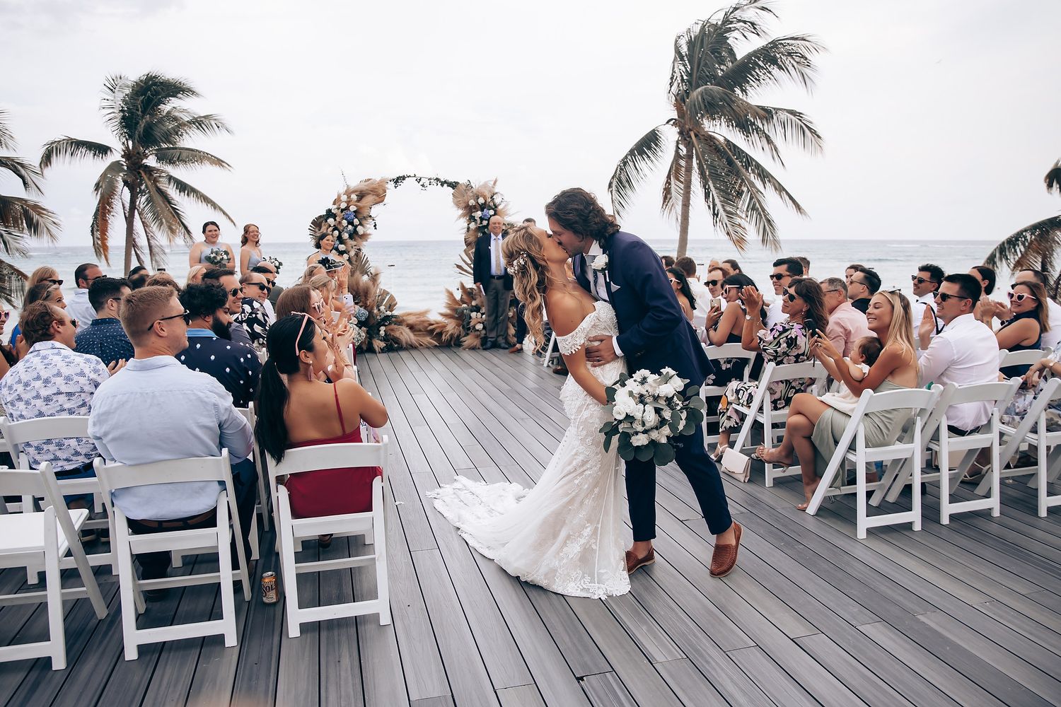 Panoramic view of a seaside wedding ceremony with rows of white chairs and tropical palm trees.