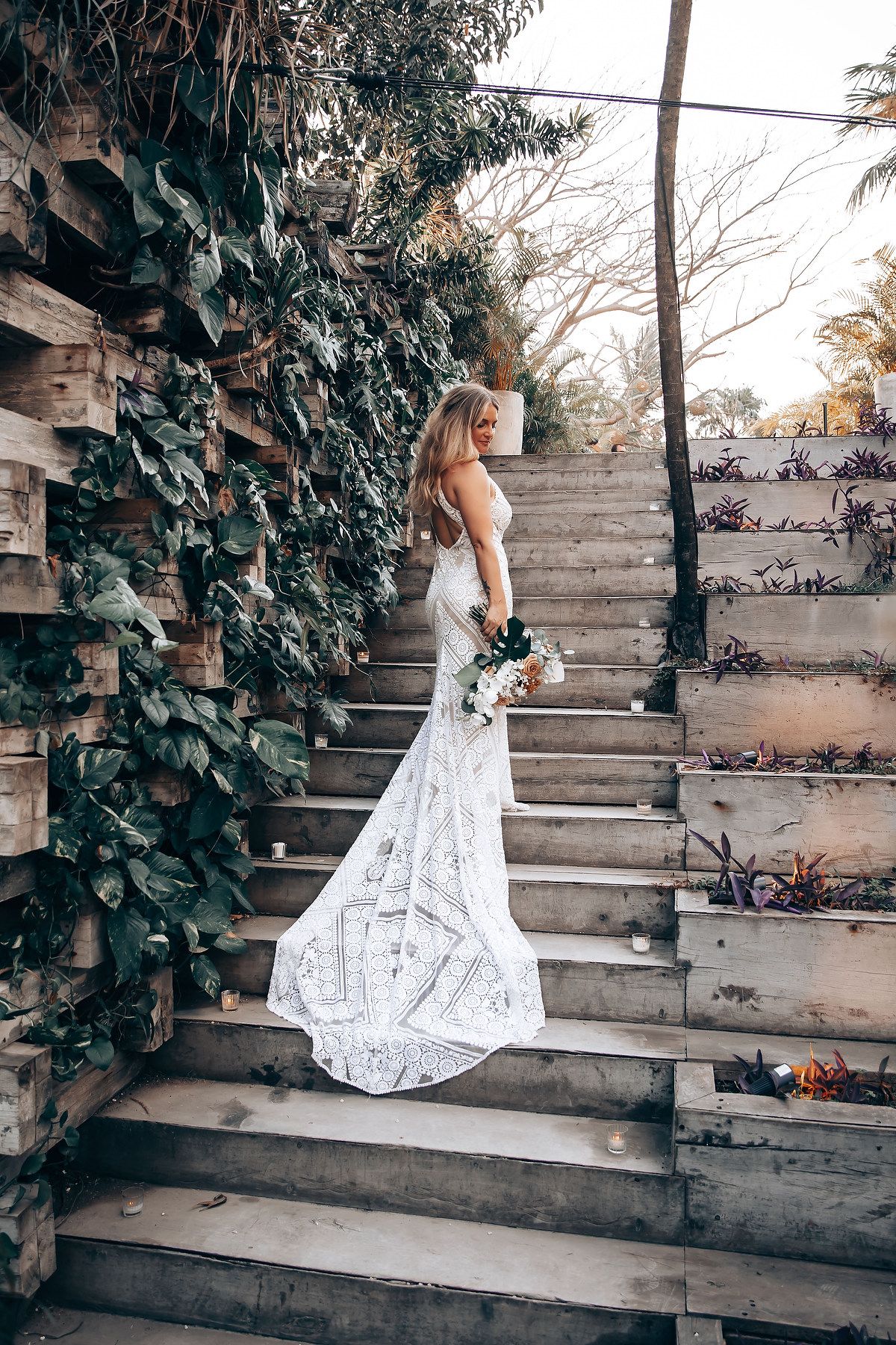 Bride in flowing lace dress descends outdoor wooden staircase surrounded by climbing vines.