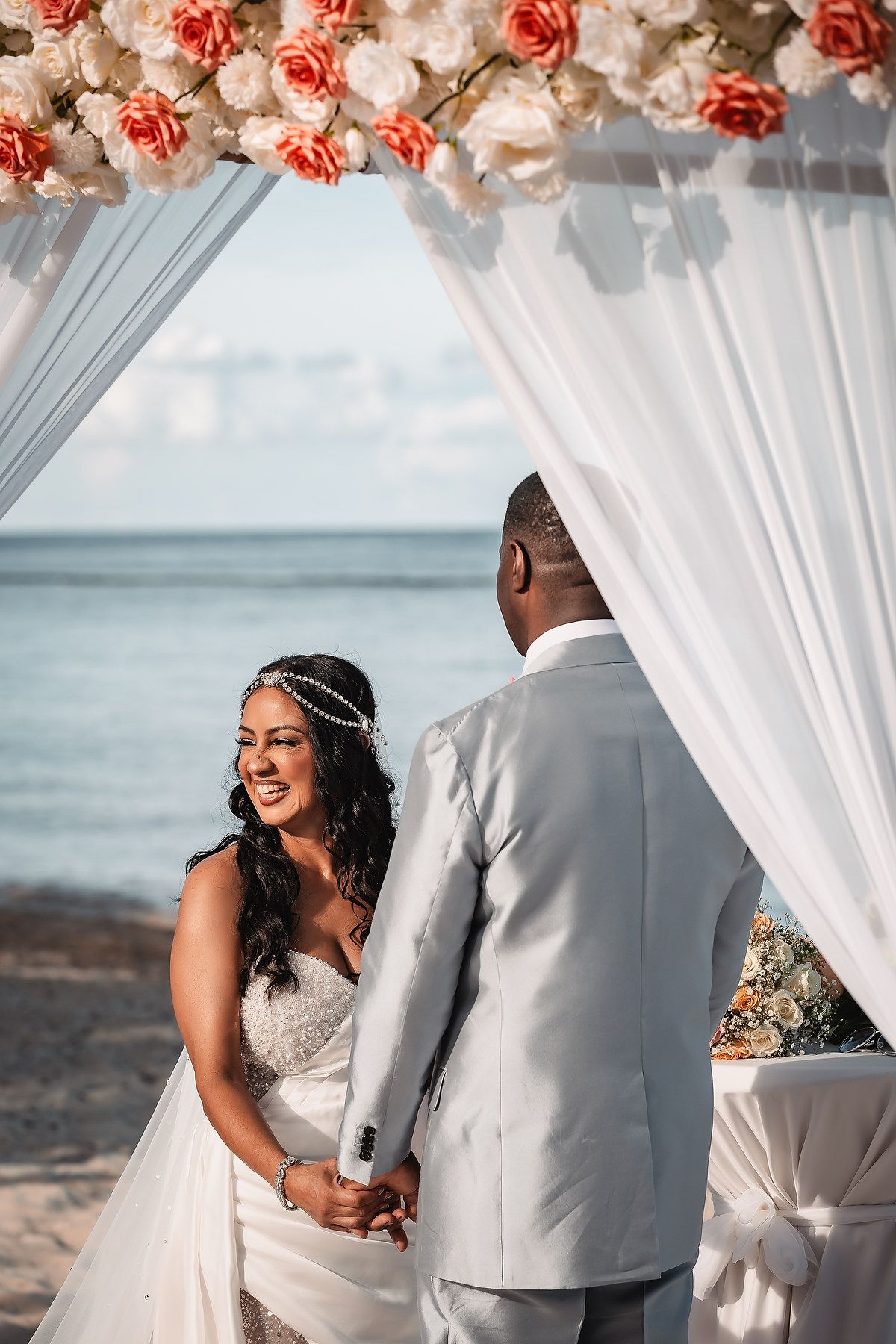 Romantic beach wedding ceremony setup with coral and white floral arch and flowing white fabric.