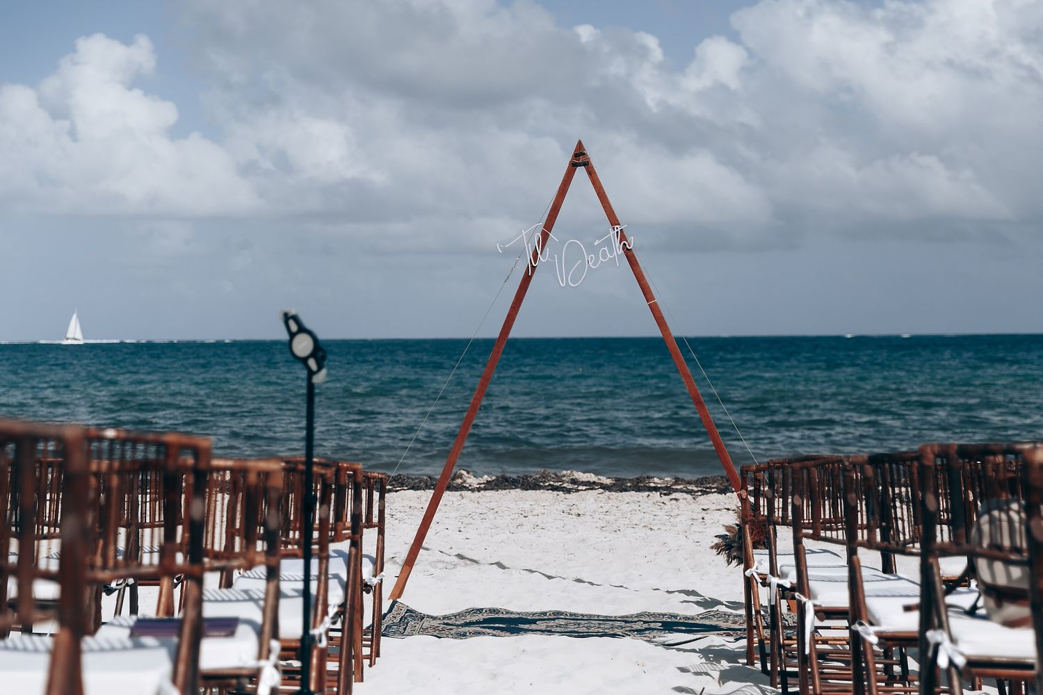 Beach wedding setup with wooden chairs and red triangular arch against blue ocean waters and cloudy sky.