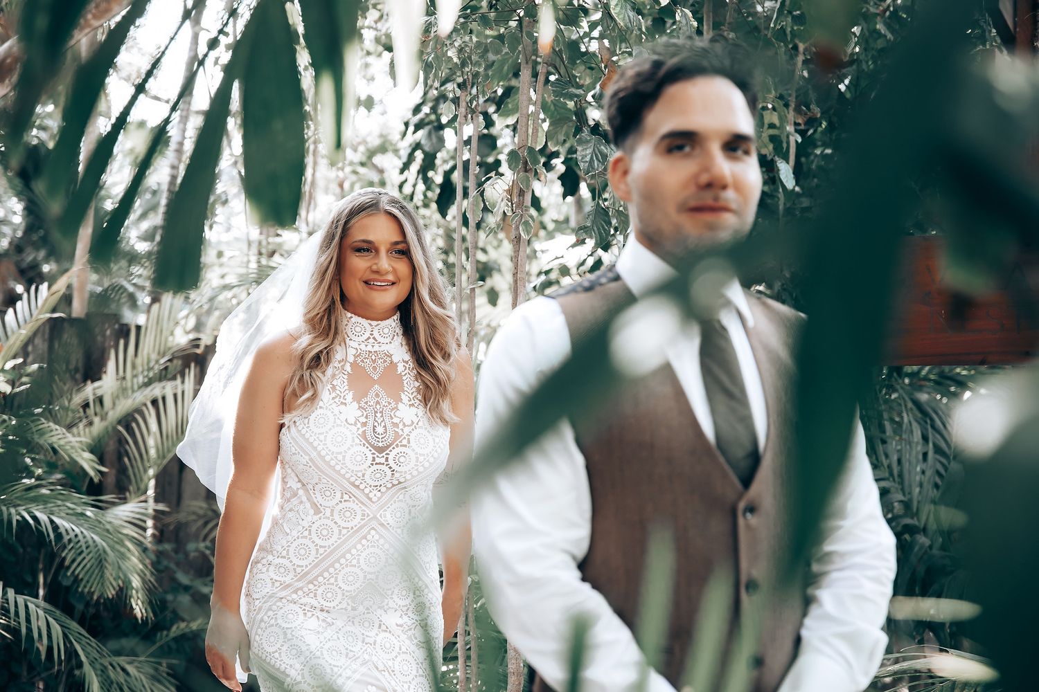 A couple in wedding attire shares a romantic moment in a lush tropical garden setting.