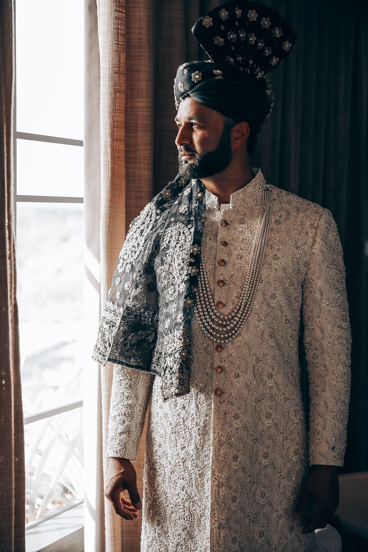 A person wearing an intricately detailed white sherwani and traditional headpiece stands by a window.