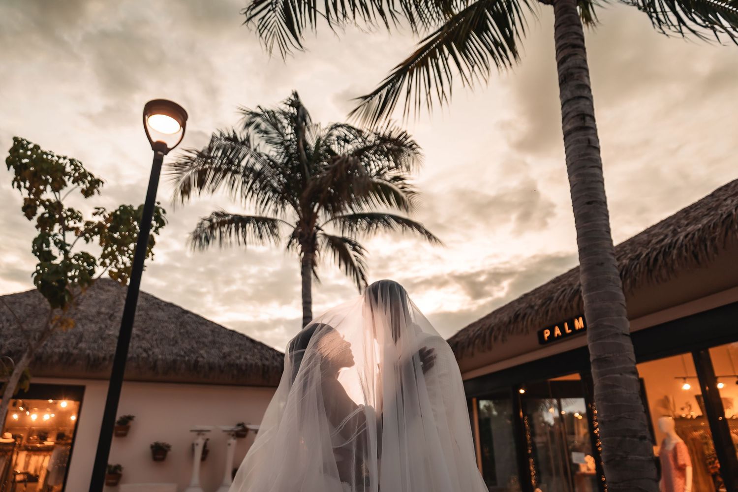 Silhouetted palm trees frame a sunset wedding scene with thatched roof buildings.