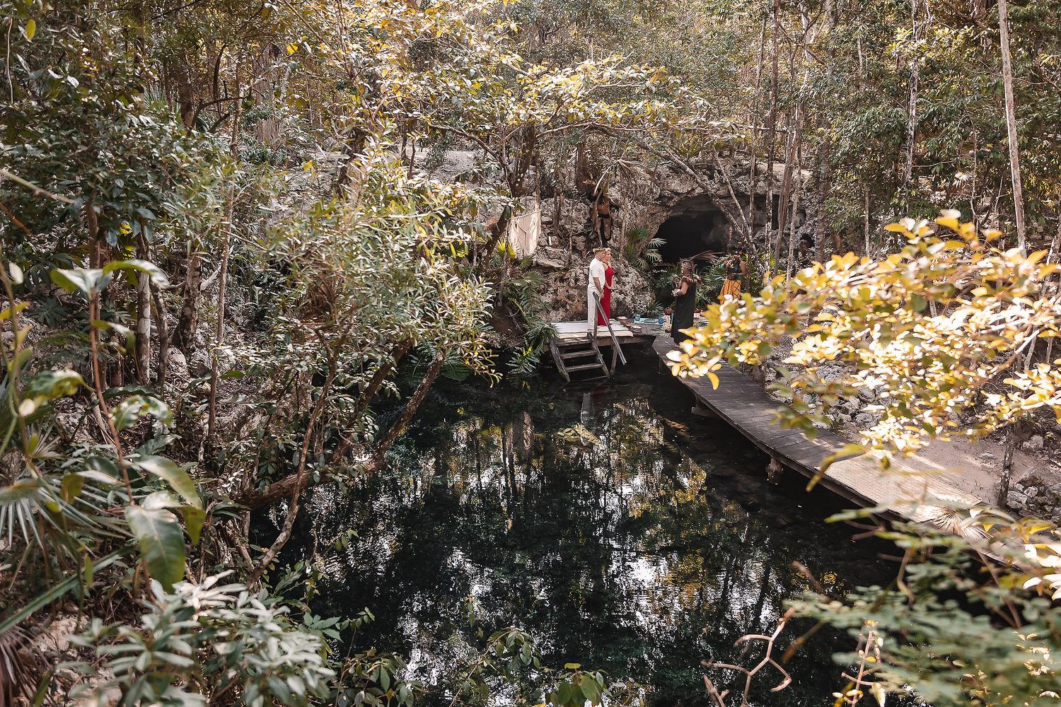 An aerial view of a wooden platform over a cenote surrounded by lush jungle vegetation.