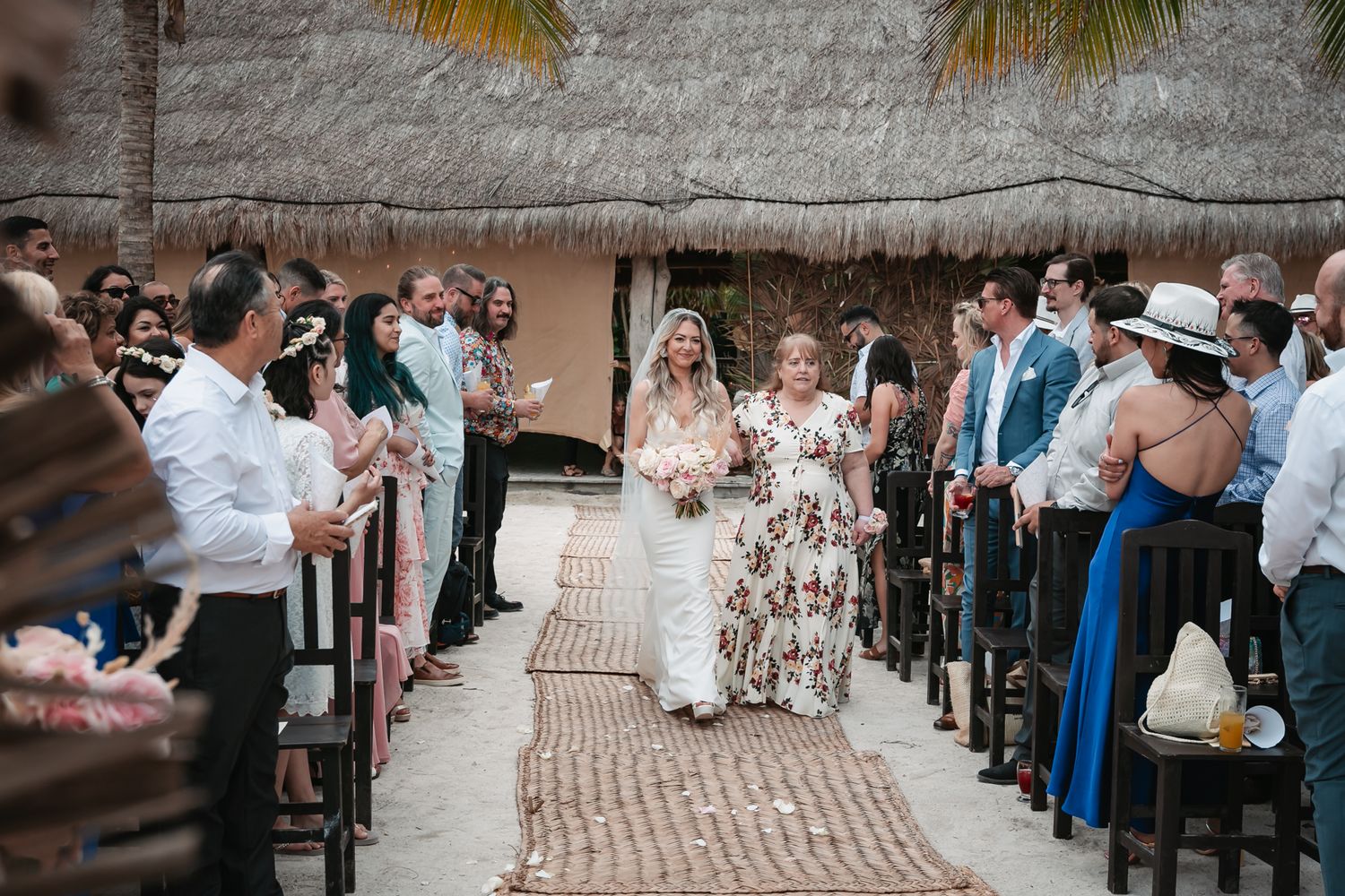 Bride walks down outdoor aisle under thatched roof with guests applauding at tropical beach wedding ceremony.