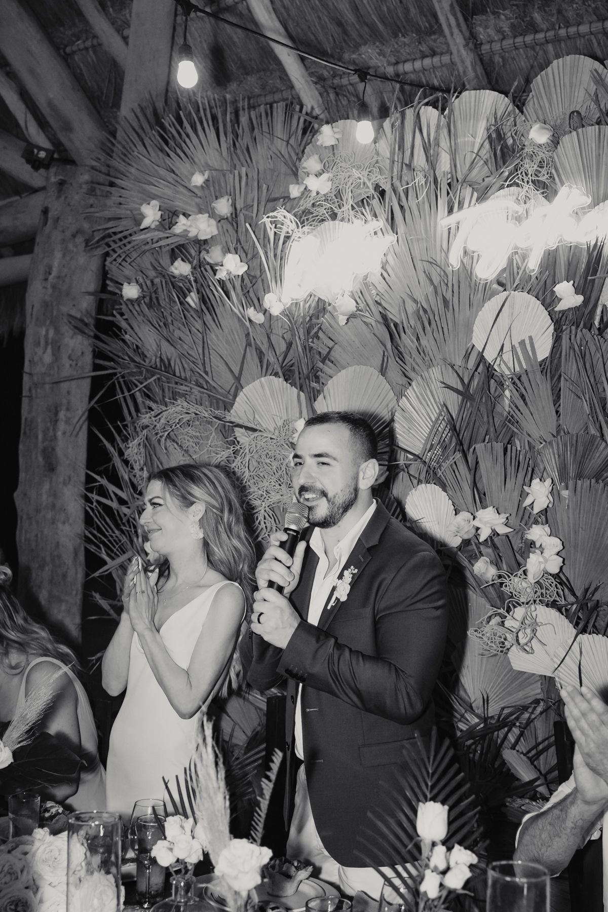 Groom speaks into microphone beside smiling bride at wedding reception table with tropical decor.