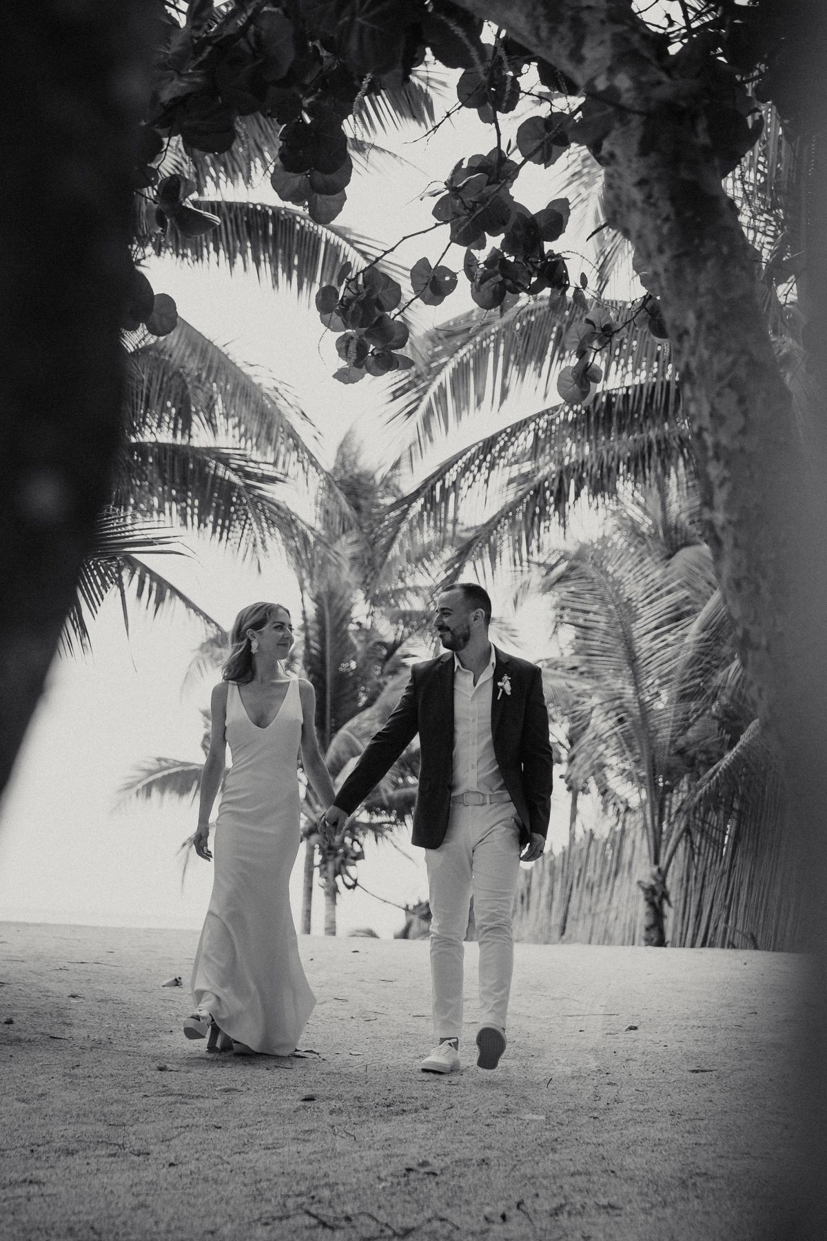 Black and white photo of bride and groom walking hand in hand under palm trees on sandy beach.