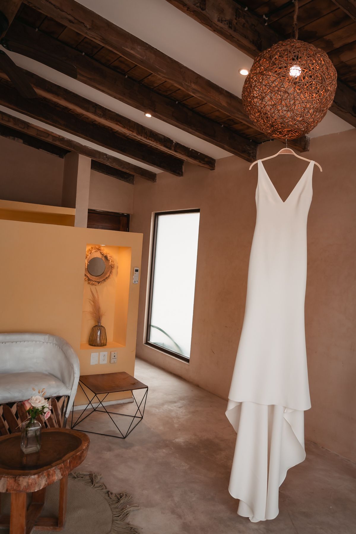 Elegant white wedding dress hanging on a hanger beneath a wicker pendant light in a rustic boho bridal suite.