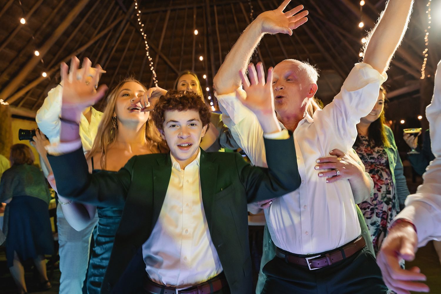 People dancing joyfully at a wedding reception in a rustic barn venue with string lights overhead.
