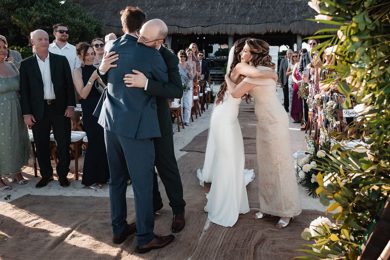 Bride and groom share emotional hugs with family members during an outdoor wedding ceremony at a tropical venue.