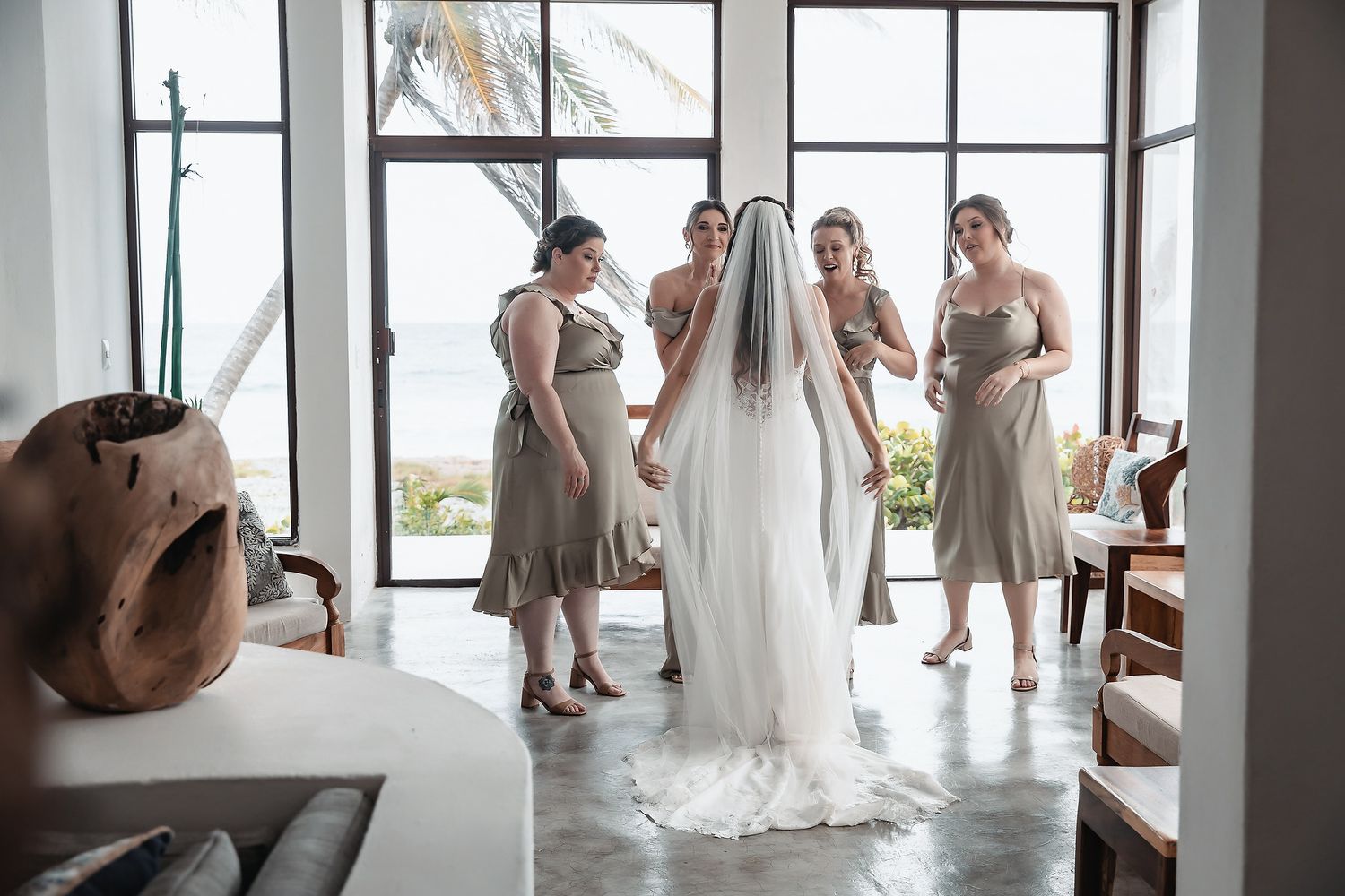 Bride in white gown and veil surrounded by bridesmaids in olive dresses indoors.