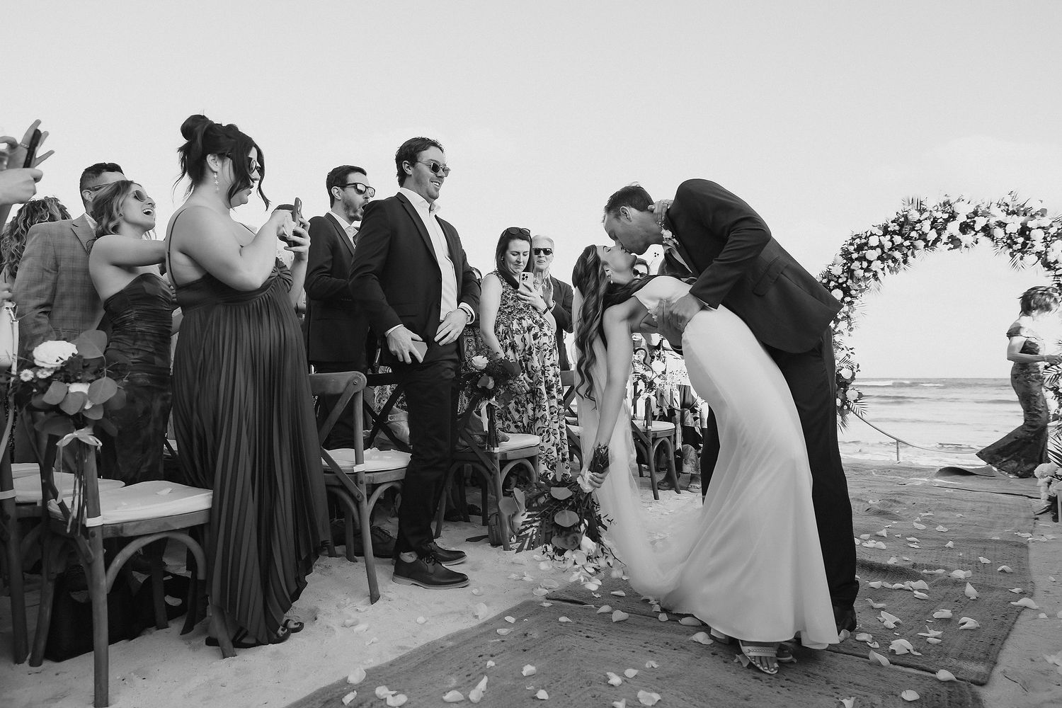 Couple sharing first kiss at outdoor beach wedding ceremony while guests celebrate in black and white photo.