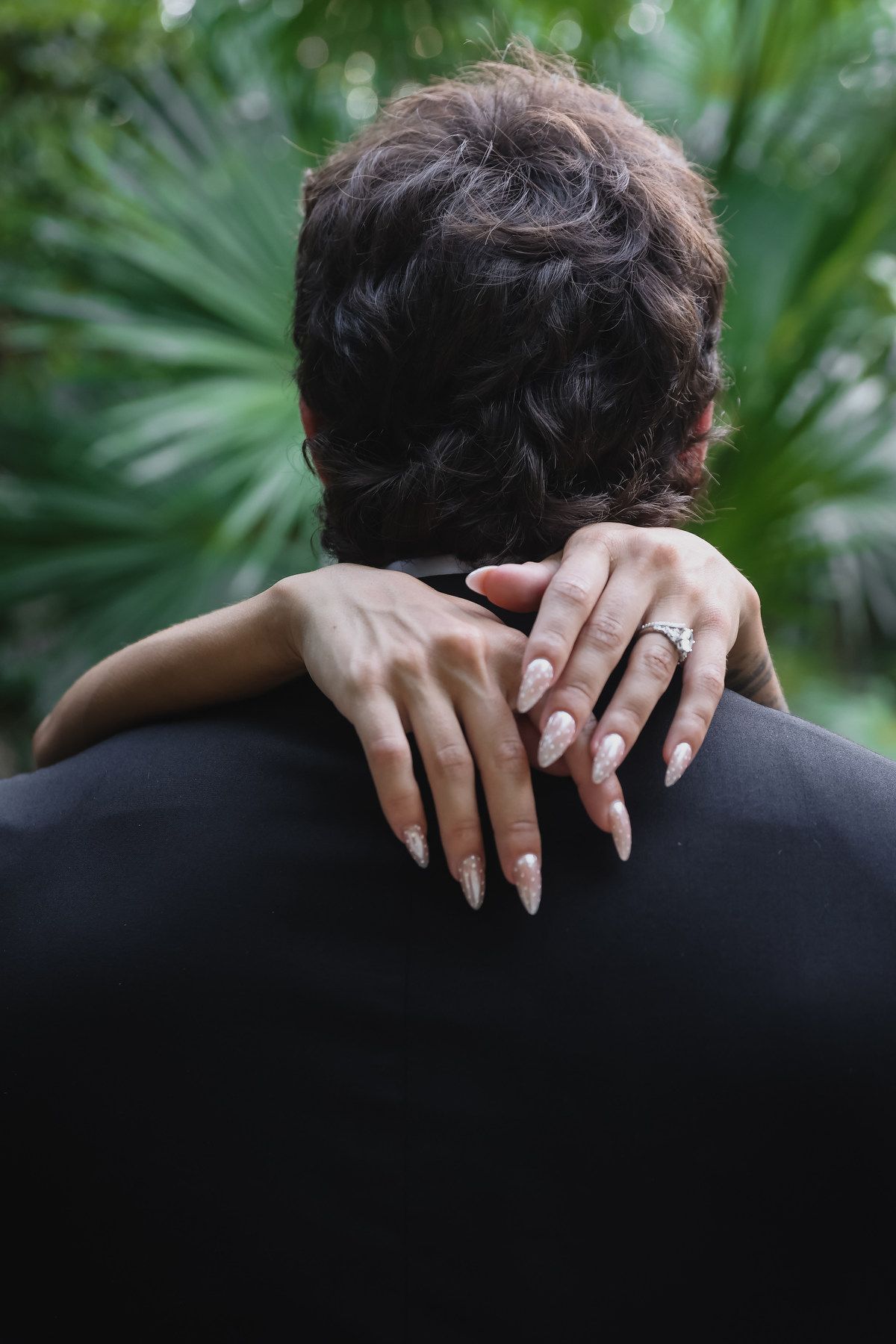 Close-up of bride's hand with diamond engagement ring resting on groom's shoulder in garden setting.