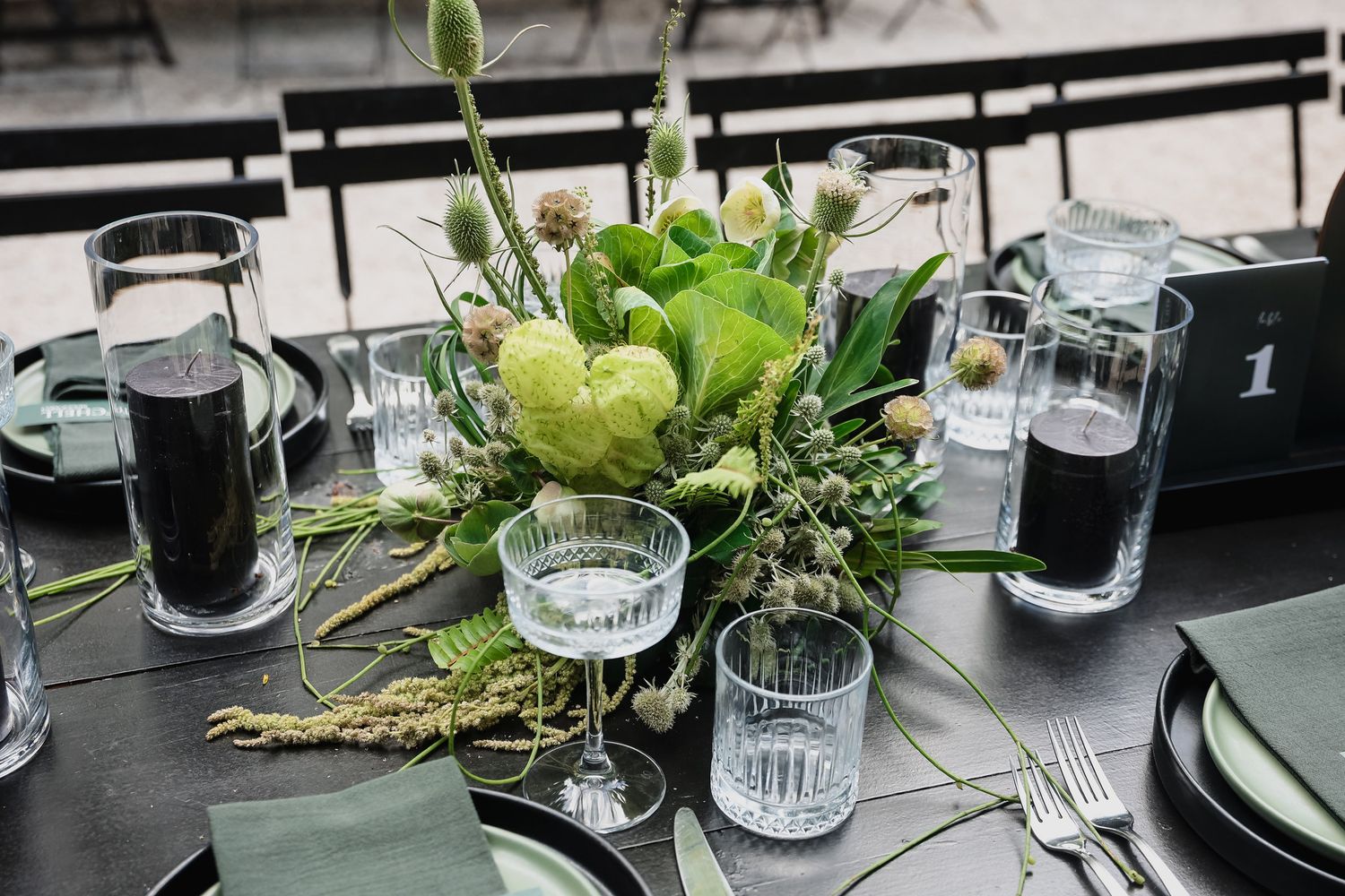 Elegant wedding table centerpiece with green florals, crystal glassware, and dark plates on a sleek black table.