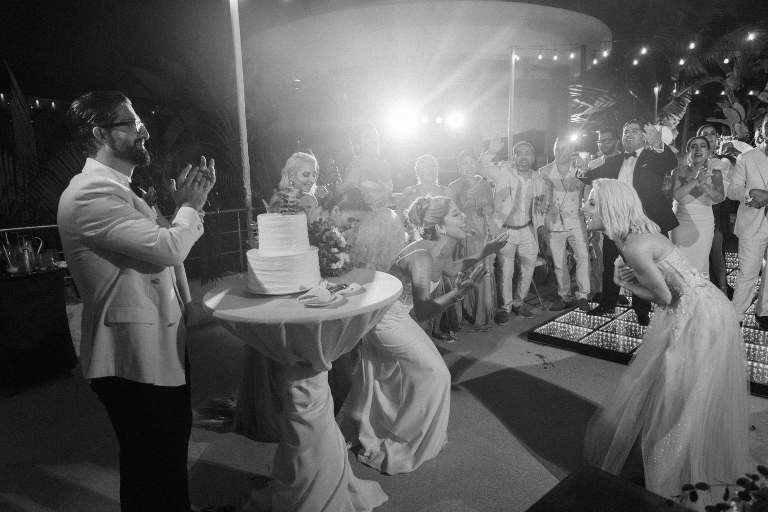 Bride and groom cutting wedding cake while guests applaud at elegant outdoor night reception.