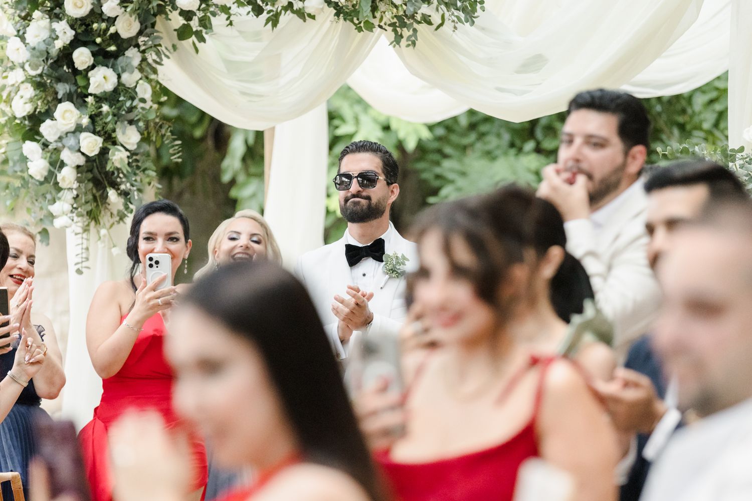 Guests watching a wedding ceremony outdoors, with a man in a tuxedo and sunglasses standing near a floral arch.