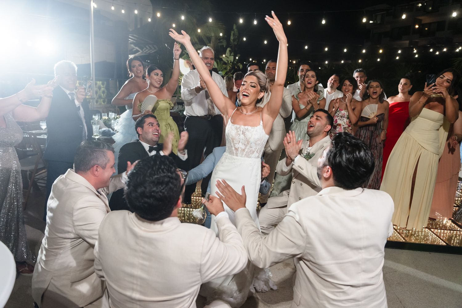 Bride in white gown celebrates joyfully at wedding reception, arms raised, surrounded by cheering guests.