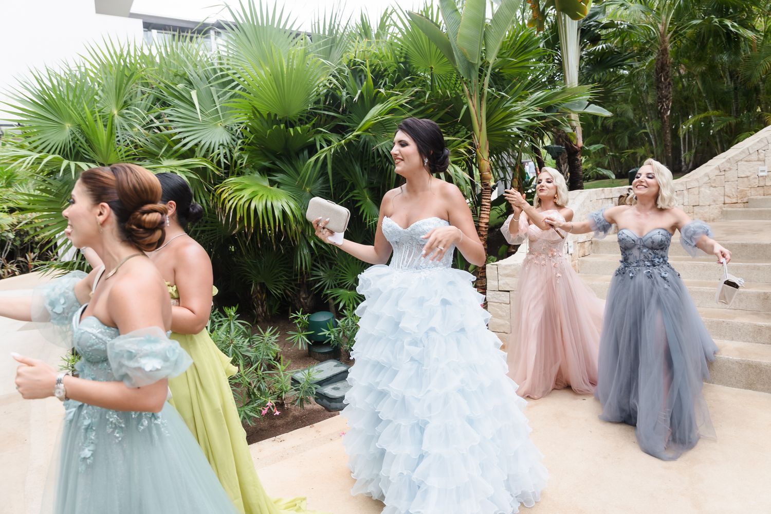 Bride in blue ruffled gown celebrates with bridesmaids in colorful dresses outdoors among tropical palm trees.