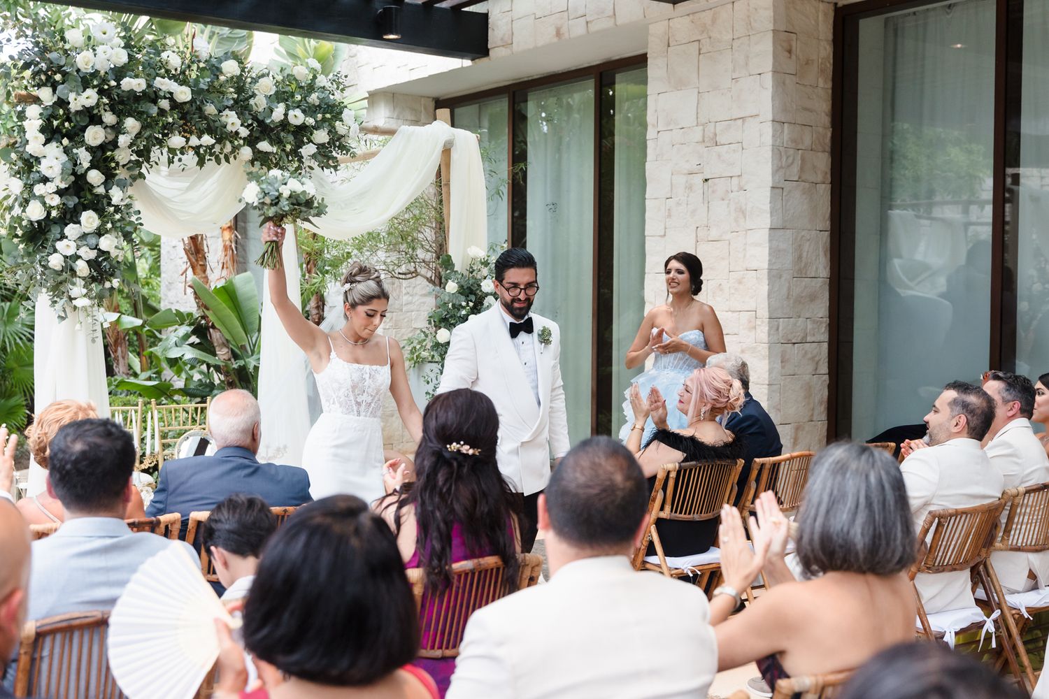 Bride and groom celebrate at outdoor wedding ceremony with guests applauding, floral arch backdrop, tropical venue setting.