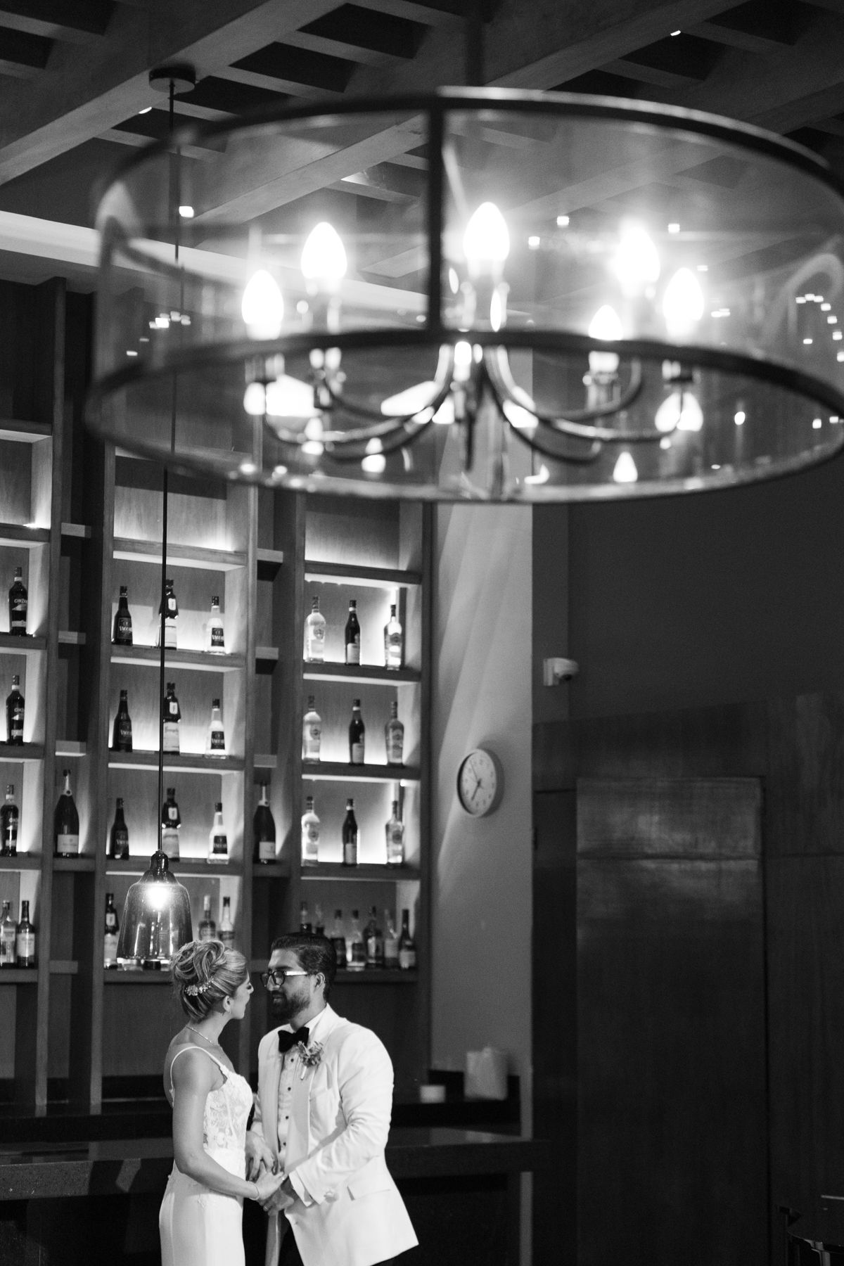 Black and white photo of bride and groom sharing a romantic moment under a chandelier at an elegant bar venue.