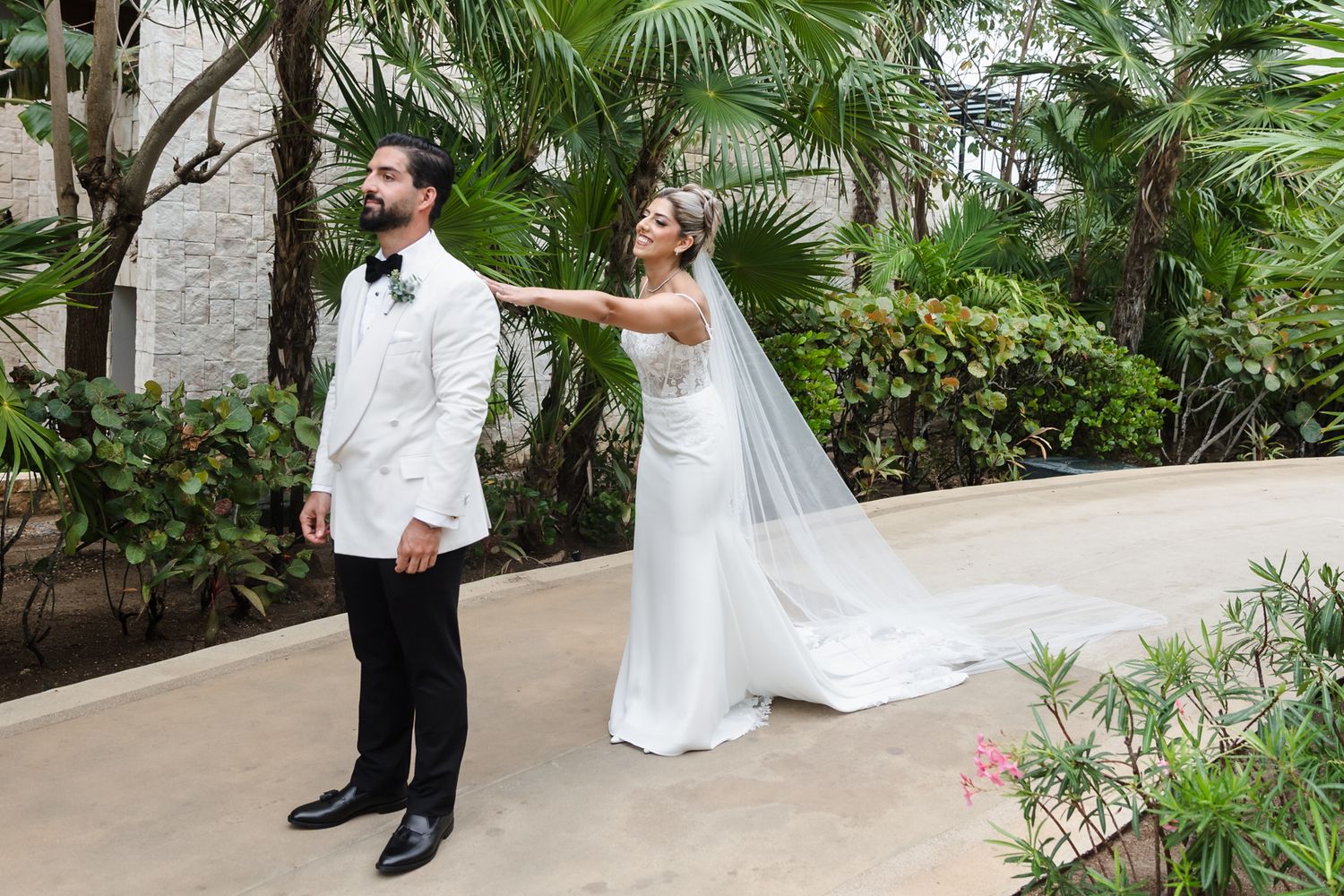 Bride in white gown and veil touches groom's shoulder during outdoor first look at tropical wedding venue.