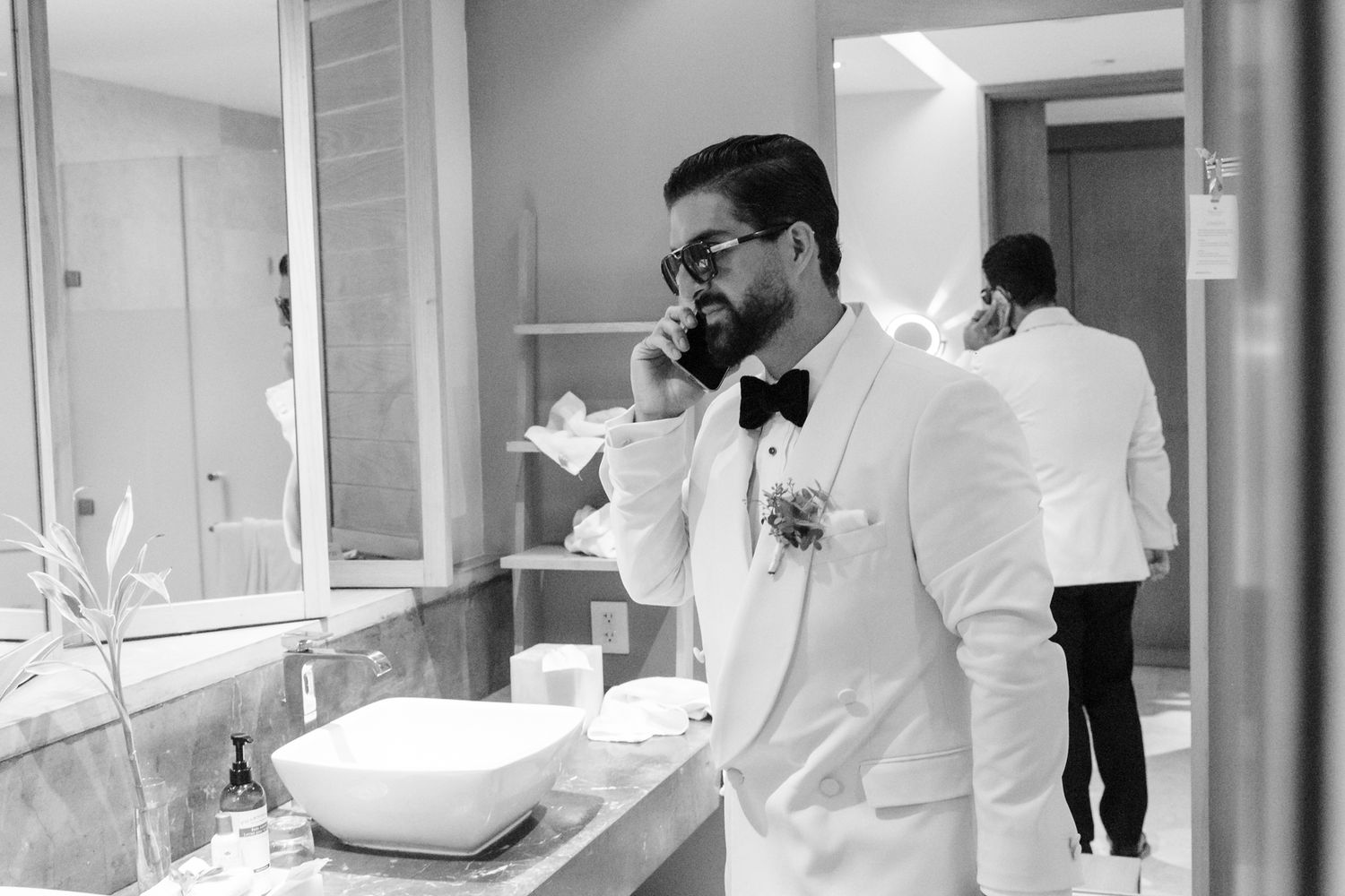 Groom in white tuxedo and bow tie talks on phone in bathroom mirror, black and white wedding photo.
