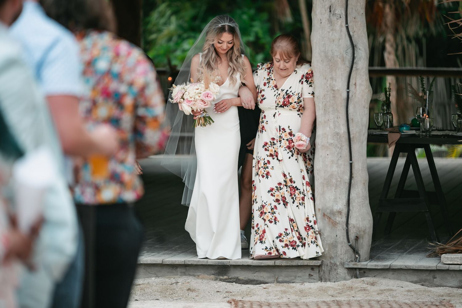 Bride in white gown and veil holding bouquet walks with guest in floral dress at outdoor wedding ceremony.