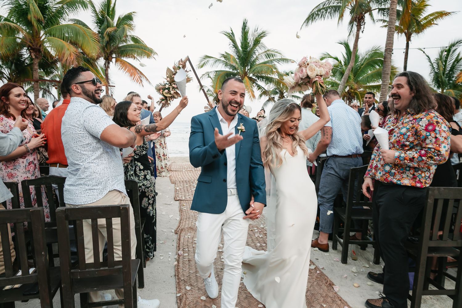 Bride and groom walk joyfully down the aisle as guests celebrate with confetti at a tropical beach wedding.