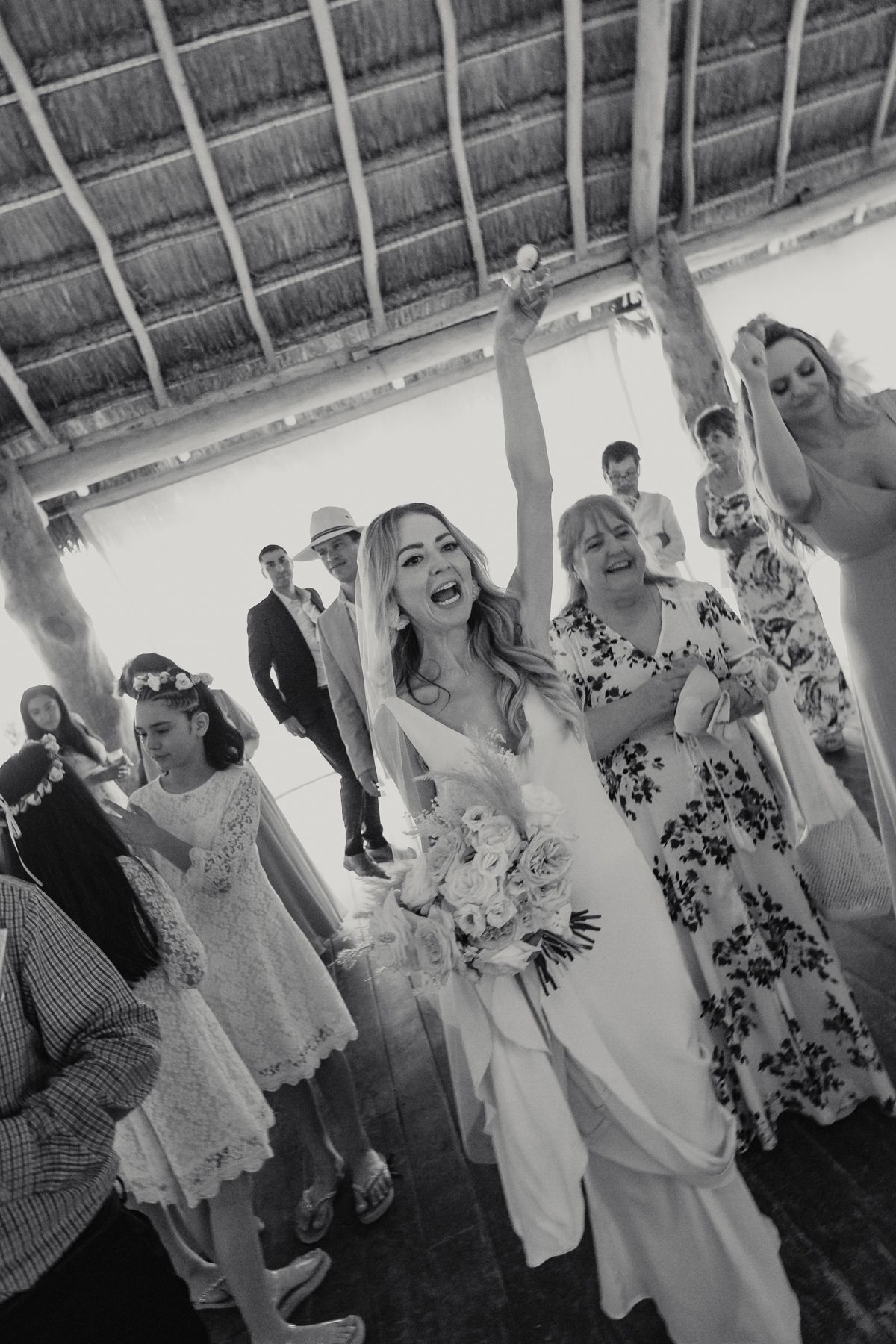 Black and white photo of bride celebrating with raised arm holding bouquet surrounded by guests after ceremony.
