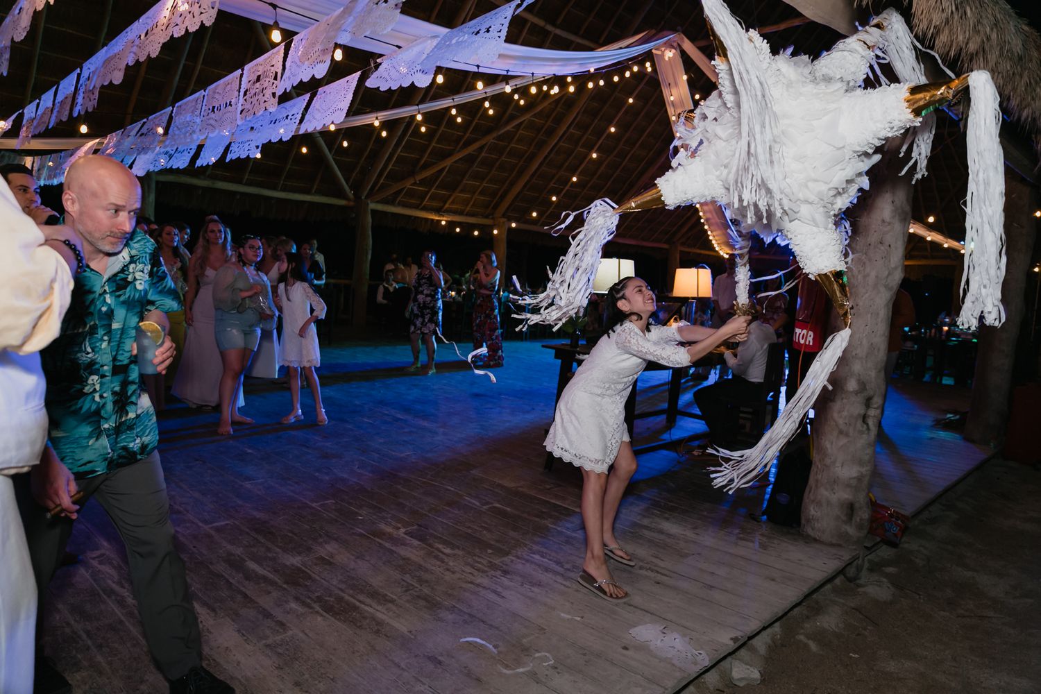 Wedding guest swings at a white pinata on the dance floor at a lively tropical beach reception.