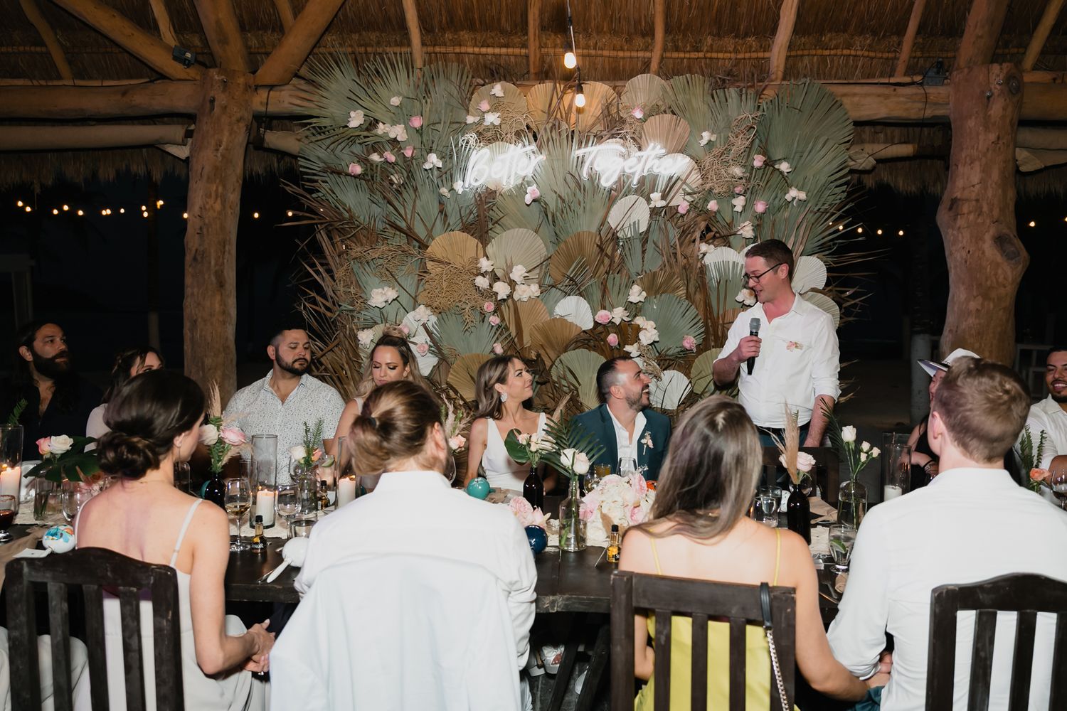 Wedding guests seated at reception dinner under dramatic floral installation in open-air tropical venue.