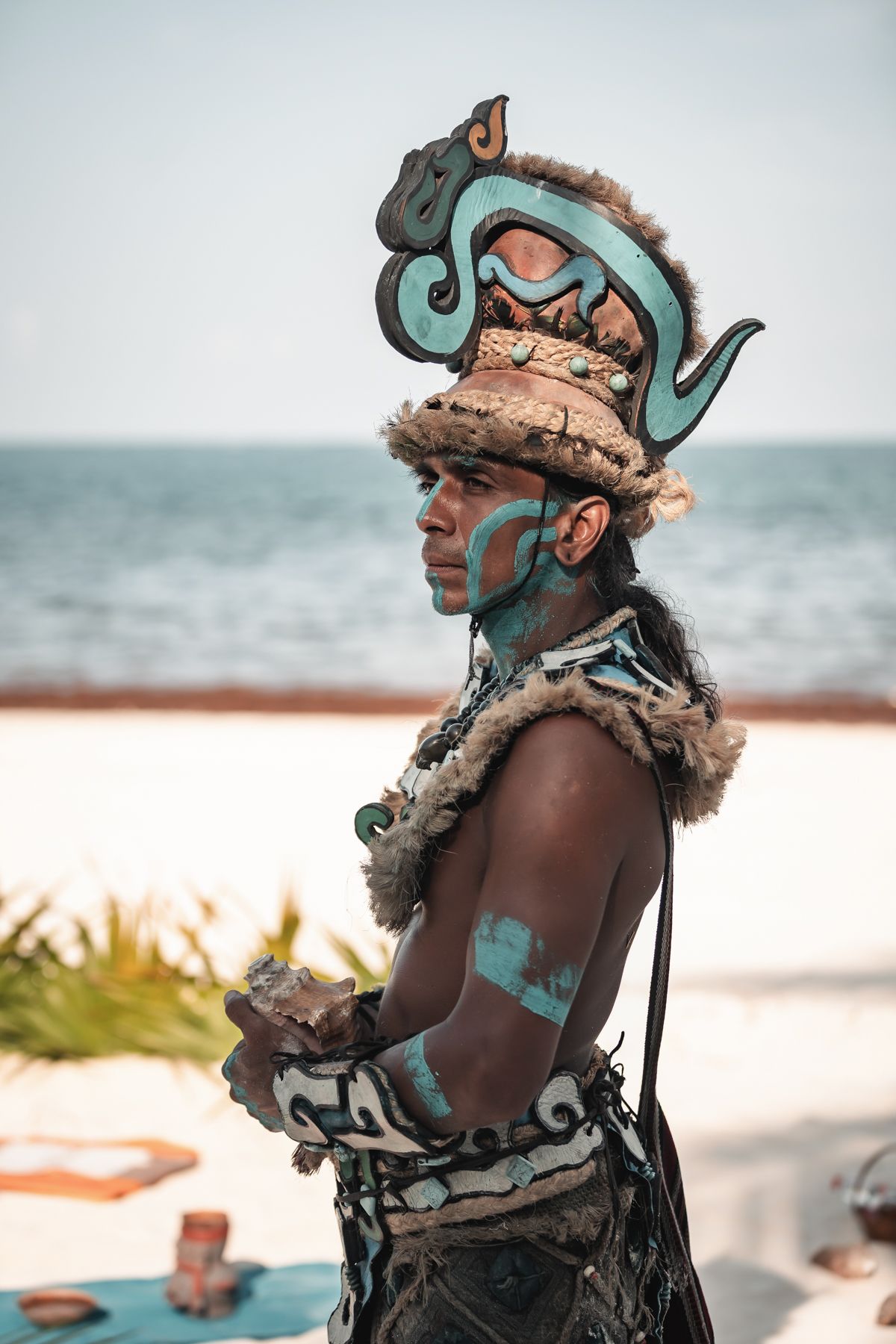 Mayan warrior in traditional turquoise body paint and ornate headdress stands on a beach in Mexico.