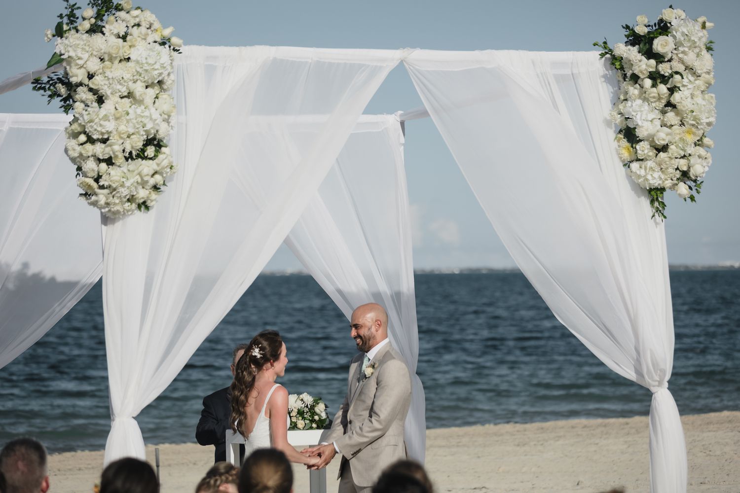 Couple exchanging vows under a white draped floral arch on a beach wedding ceremony by the ocean.