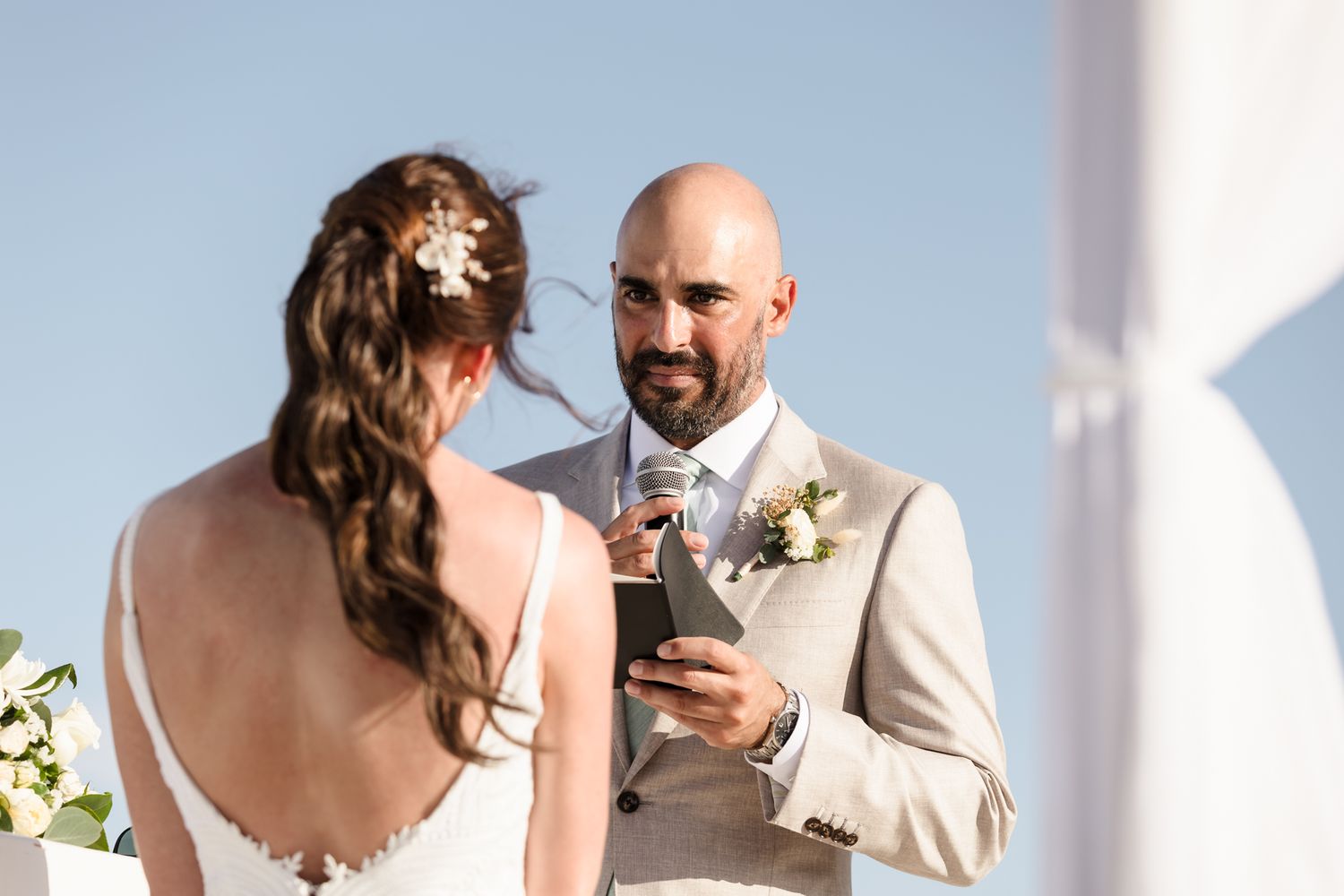 Bride and groom exchanging vows outdoors, groom in beige suit with boutonniere reading from phone under blue sky.
