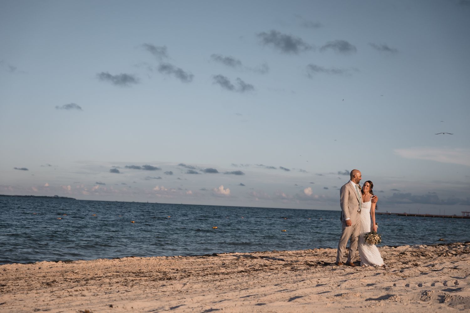 Wedding couple walking hand in hand on a sandy beach with calm blue ocean at dusk.